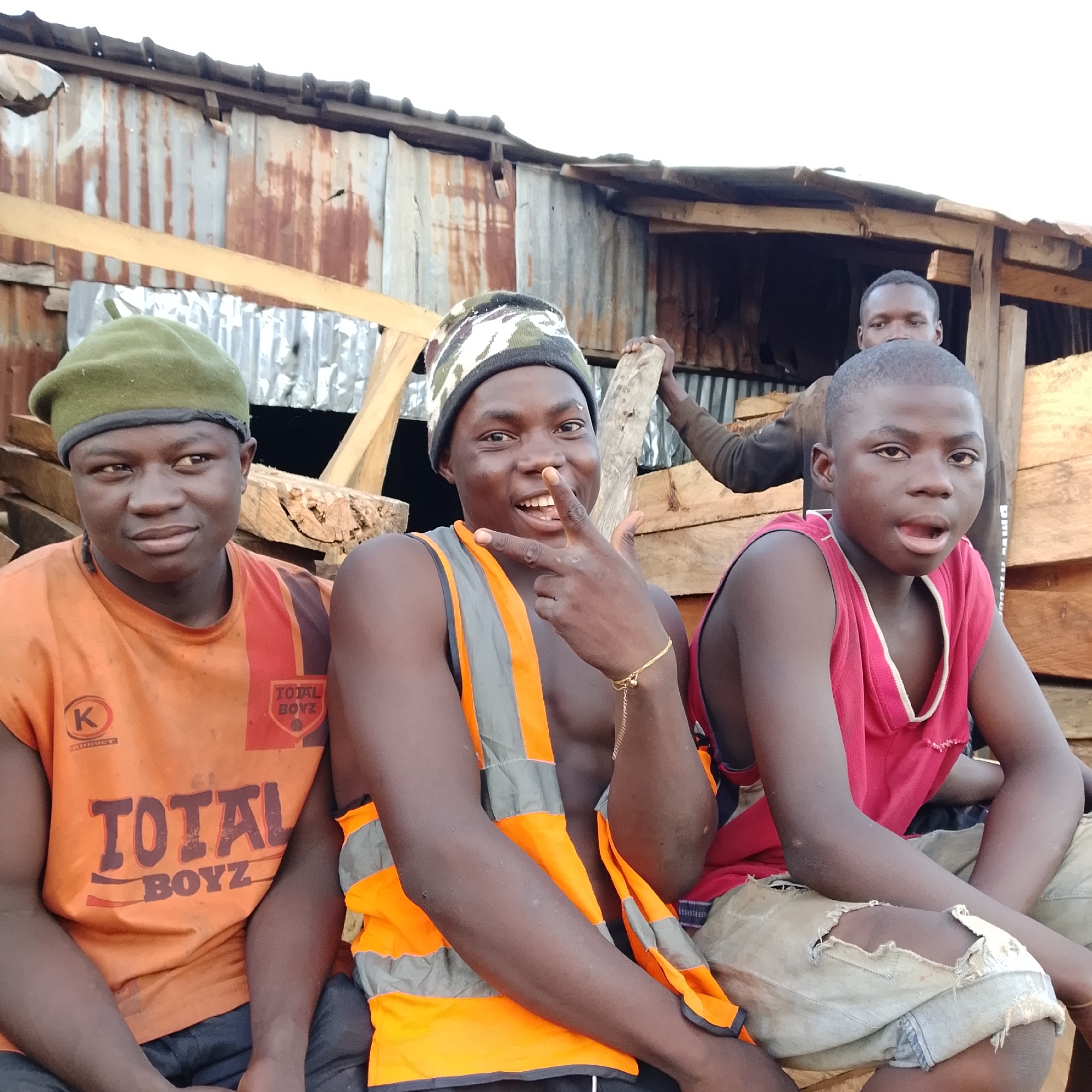 A group of four young males are posing for a photograph, with three of them in the foreground and one partially visible in the background. The setting appears to be an outdoor construction site, evidenced by the corrugated metal and wooden structures in the background, which are weathered and appear makeshift.
In the center foreground, a young male wearing a camouflage beanie and a reflective safety vest over his bare chest smiles and makes a "rock on" hand gesture. To his left, a young male in a green beanie and an orange t-shirt with "TOTAL BOYZ" printed on it looks to the side and smiles slightly. To the right of the central figure, another young male in a red tank top and ripped light-colored pants stares directly at the camera with his mouth slightly open. In the background, a fourth young male is visible, leaning against a wooden beam and holding a piece of wood.
The lighting suggests it is daytime, likely overcast given the even illumination. The overall impression is one of camaraderie and youthful energy amidst a utilitarian environment. The location is not specified beyond "Unknown, Unknown."
