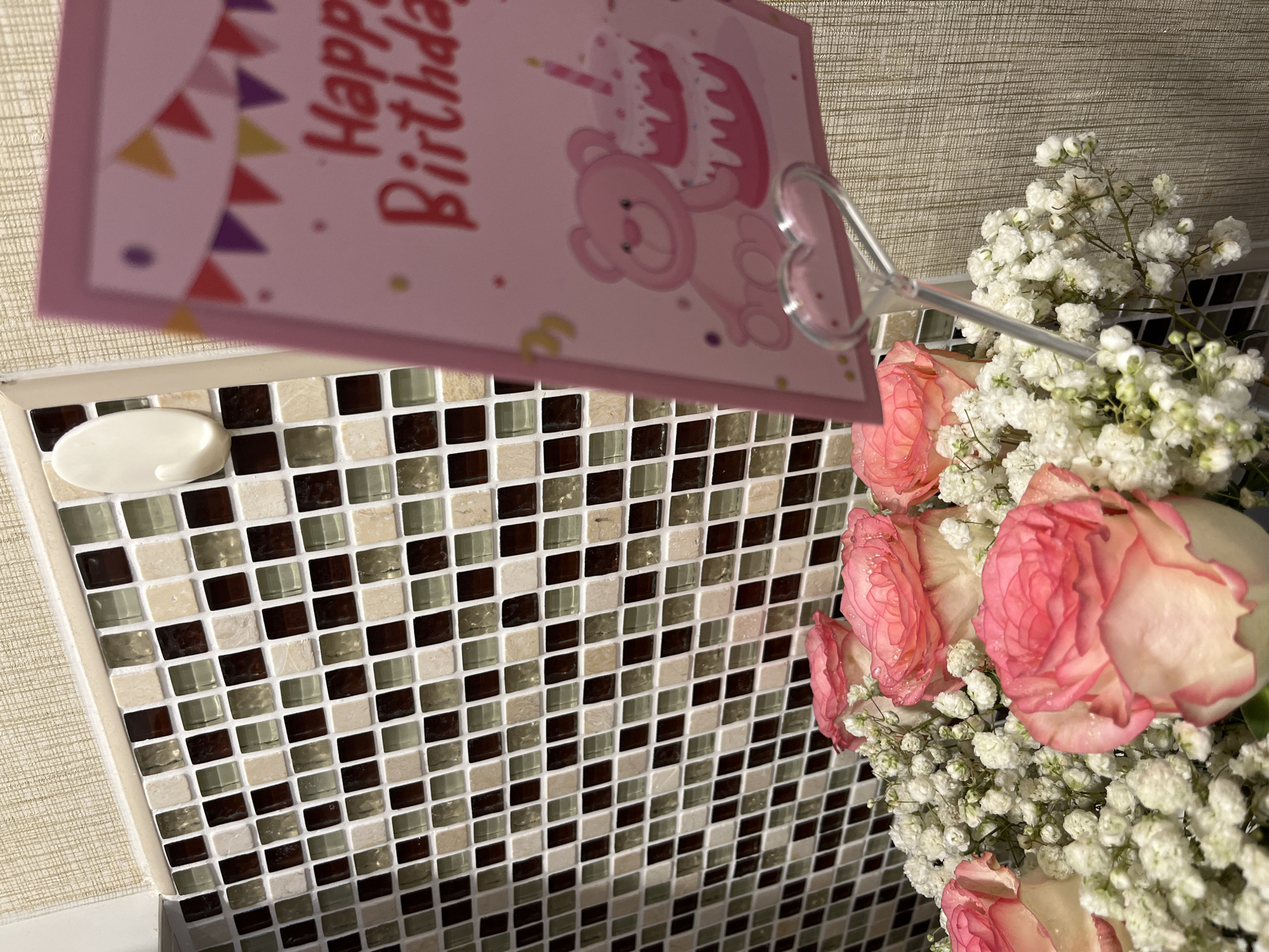 The image features a close-up view of a bouquet of pink roses and white baby's breath flowers, accompanied by a pink "Happy Birthday" card with a cartoon bear holding a cake. The card is displayed using a heart-shaped holder. The background consists of a tiled wall with a mosaic pattern of dark brown, beige, and white square tiles. A white soap dish is visible on the upper left corner of the tile. The setting is unspecified, but the context of a birthday celebration is evident. The image appears to be indoors. The location of Pattaya City, Thailand, is extraneous and not discernable from the visual information.
