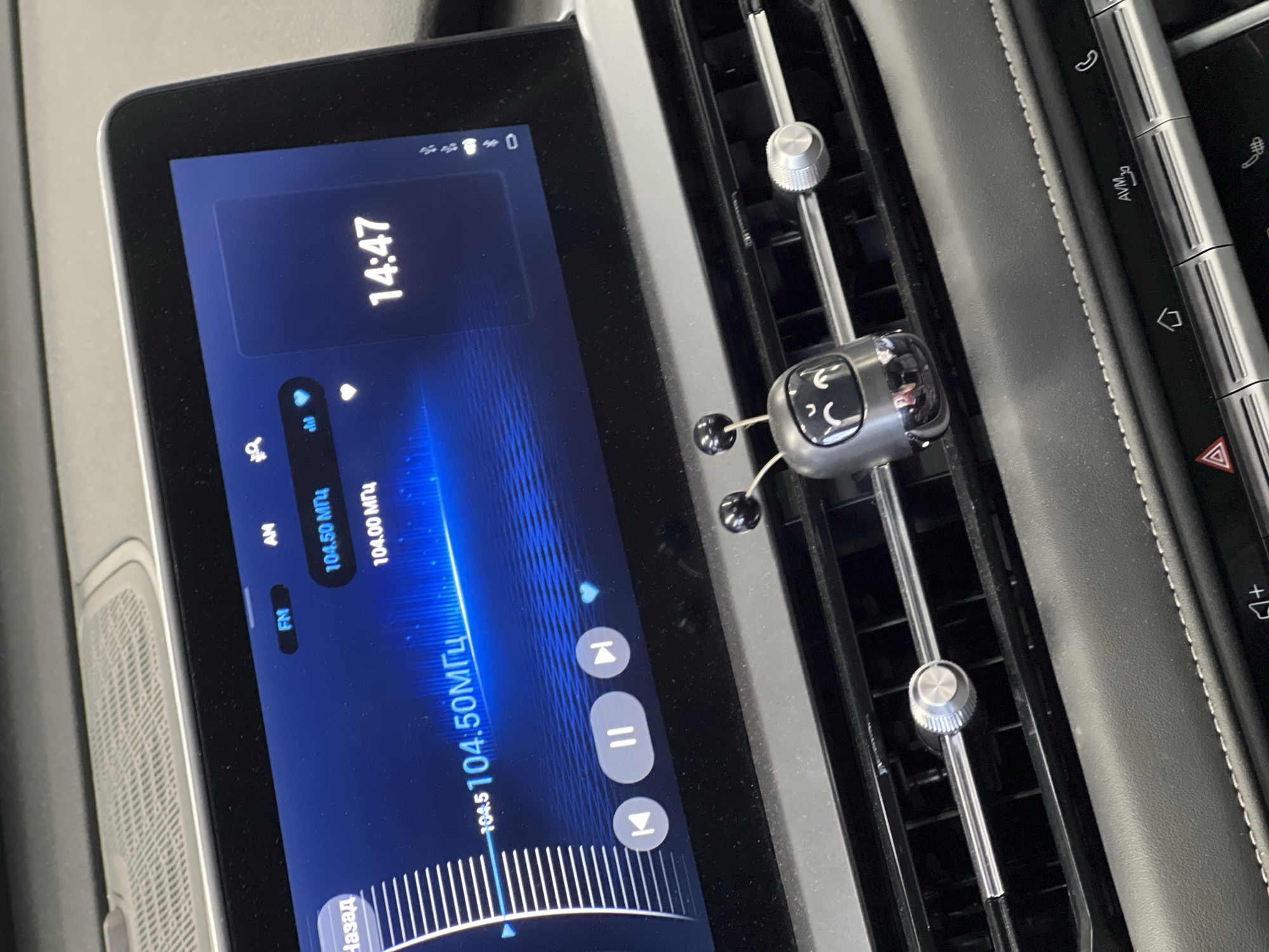 The interior of a vehicle in the city of Izhevsk, Russia, is depicted, centered on the infotainment screen and dashboard. The digital infotainment screen displays a radio interface set to 104.50 МГц (MHz), with 104.00 МГц visible as an alternative preset. The current time is 14:47. Playback controls, including previous, pause, and next track/station, are visible at the bottom of the screen. Below the screen are horizontal air vents, to which a small, dark grey, robot-like air freshener or ornament with black antennae is affixed. A circular, silver knob for adjusting airflow is integrated into the air vent's central slat. Below the air vents, a padded dashboard segment with white stitching is visible, followed by a row of physical buttons including icons for hazard lights, home, AVM (Around View Monitor), and a telephone.