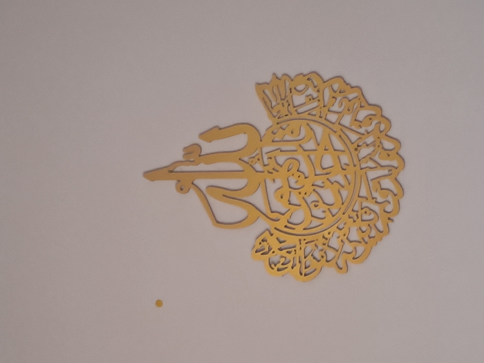 The scene depicts an indoor environment with a light grey wall. Centrally mounted on the wall is a golden, ornate decoration featuring Arabic calligraphy. The upper portion of the decoration forms the stylized word "Allah," while the lower part consists of a circular, filigree-like design with additional intricate Arabic text and floral patterns. Approximately 15cm above and 10cm to the left of the decoration's top-left edge, a small, circular, yellow-orange mark, approximately 2mm in diameter, is visible on the wall. No individuals are present. The location is Undeterminable, city of Unknown, Unknown.