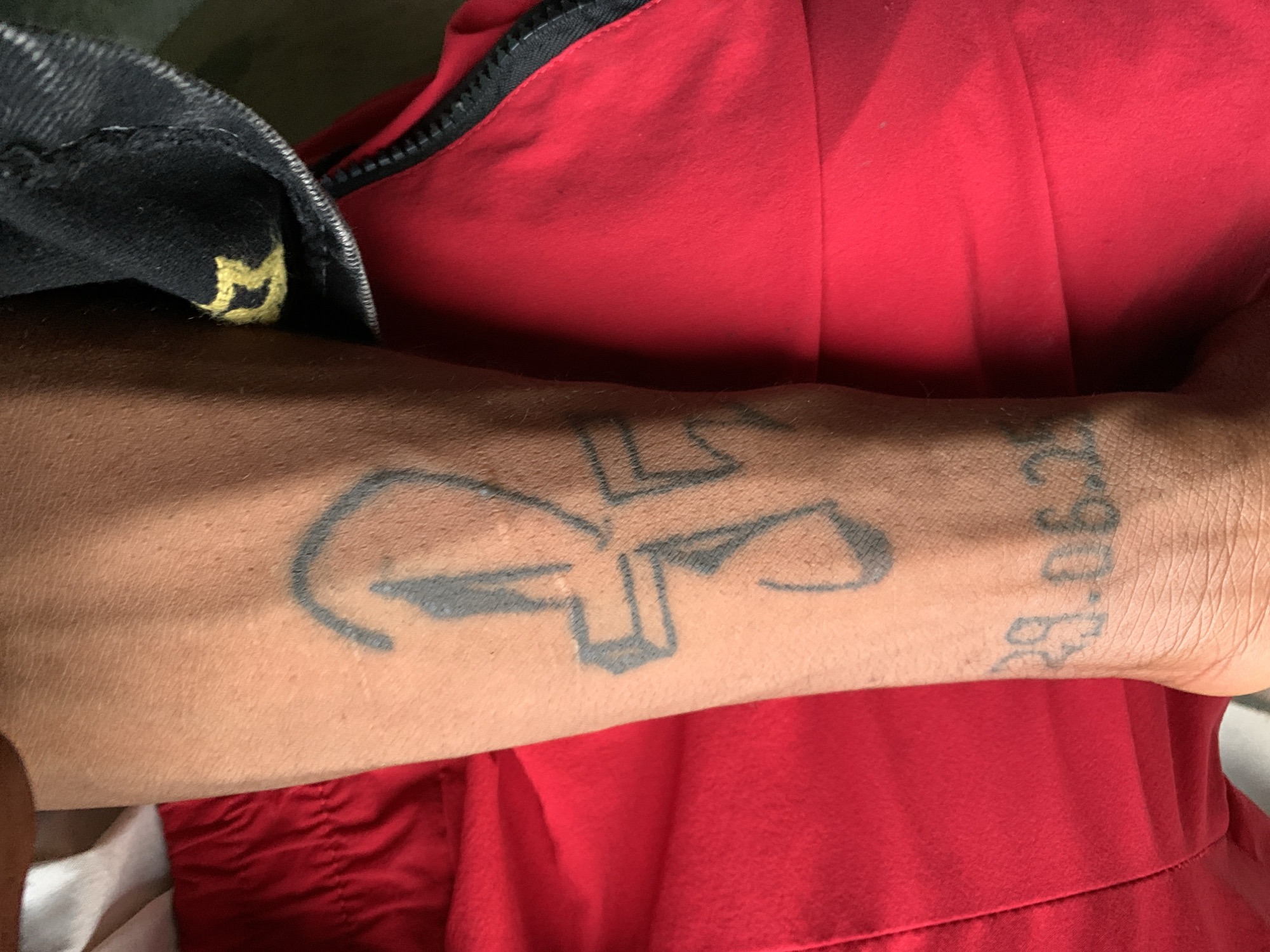 The image shows a person's forearm with two tattoos. The first tattoo appears to be an anchor and a cross combined. The second tattoo is a date, "29.06.68". The person is wearing a red garment and a black denim jacket with a yellow detail. The location is not provided in the image.