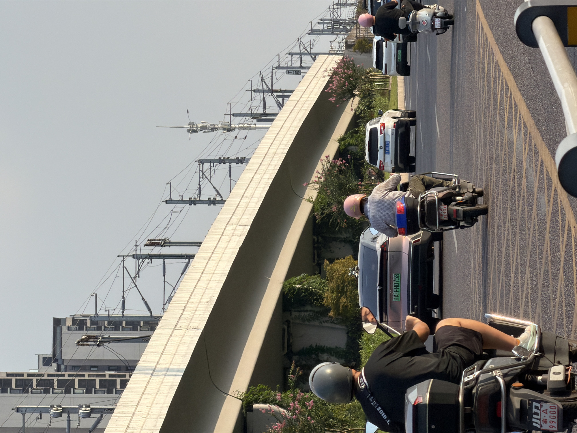In Jiangjin, China, an outdoor daytime scene unfolds with traffic flowing under an elevated highway and railway infrastructure. In the foreground, the back of a person on a scooter is visible, wearing a black t-shirt and shorts, with a grey helmet. To their right, another person on a scooter, wearing a patterned shirt and a pink helmet, is also visible. In the middle ground, two white SUVs are driving on the road, and behind them, a silver car is seen with its license plate readable as "A FH0758". The road is paved with asphalt and marked with yellow lines. The elevated structure above is made of concrete and supports overhead power lines for trains. Green vines grow along the supports of the elevated structure. The sky is clear and pale blue. The image captures a snapshot of urban transit and infrastructure in Jiangjin.
