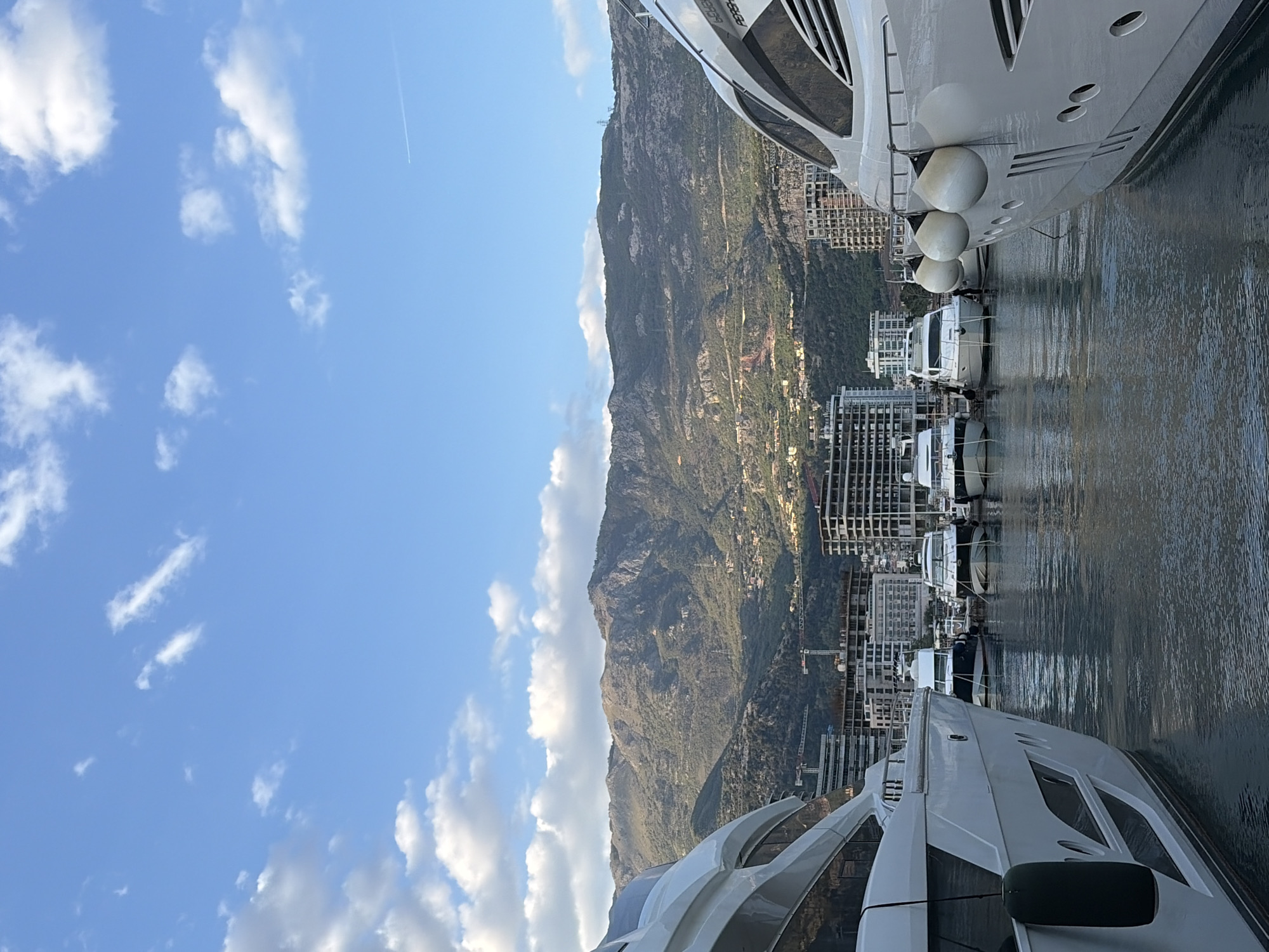The image, originally rotated ninety degrees clockwise, presents a vibrant marina scene in Budva, Montenegro. Large, luxurious white yachts dominate the foreground, flanked by calm, dark water reflecting their forms. On the right side, a prominent yacht displays its sleek hull, adorned with several spherical white fenders and "AZIMUT" clearly visible in white text on its side. To the left, another large yacht is partially visible, showcasing modern architecture with dark windows and a dark, rectangular element.
Between these prominent vessels, several smaller boats are moored, contributing to the active marina atmosphere. The water, gently rippled, suggests a serene environment.
In the midground, numerous multi-story buildings, likely hotels or residential complexes, line the waterfront and ascend the lower slopes of the dramatic, verdant mountain range that forms the backdrop. These buildings appear contemporary in style. The mountains themselves are densely covered in green vegetation, with roads and scattered structures visible on their slopes.
Above, the sky is a bright, clear blue, dotted with scattered white, fluffy clouds. A faint, thin white contrail from an airplane stretches across the upper right sky. The bright illumination indicates either late morning or early afternoon. The scene conveys a sense of luxury, natural beauty, and a popular coastal resort destination.