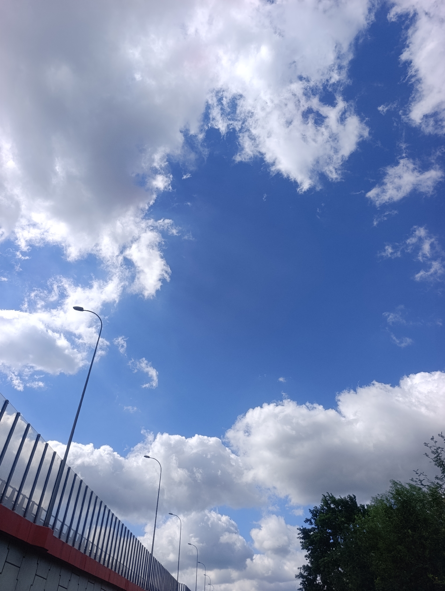 This is an outdoor, eye-level, vertical, low-angle shot of a bright blue sky filled with white and gray clouds. In the lower left quadrant, a red-and-gray sound barrier wall runs diagonally upwards, with a series of vertical, reflective metal slats and supports. Several streetlights are visible along the wall, extending upwards into the sky. In the far right of the image, the tops of dark green trees are visible against the sky. The scene appears to be a section of a highway or elevated road in Krakow, Poland, likely near a residential or urban area given the presence of the sound barrier and trees. No people or animals are present.