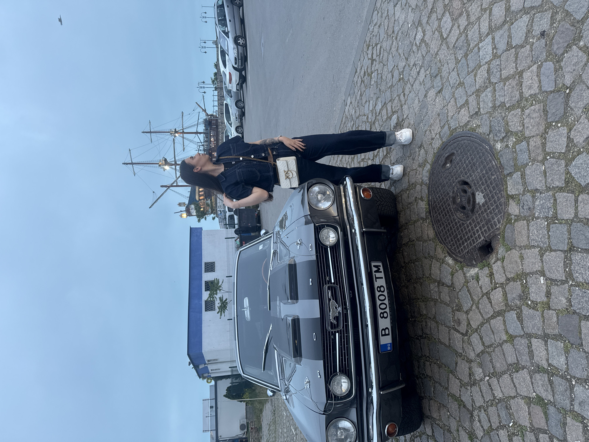 The image depicts an outdoor scene in the city of Varna, Bulgaria, during an overcast day or at dusk. In the foreground, a gray 1965-1966 Ford Mustang classic car with dual gray racing stripes on its hood is parked on a cobblestone surface. The car's front features circular headlights, a chrome bumper, and a prominent hood scoop. The license plate, "B 8008 TM," with a "BG" country identifier, is affixed to the front bumper. To the right of the car, an adult female stands, facing right and gazing slightly upward and rightward. She has long dark hair and is dressed in a dark denim short-sleeved top, dark denim wide-leg trousers cuffed at the ankles, and white sneakers. She carries a small, light-colored handbag with a gold chain strap across her body. Her left hand is raised towards her hair.
The ground is composed of gray cobblestones with sparse green vegetation, and a dark circular metal manhole cover is visible in the lower right. In the midground, behind the car and to the left, a building with a white base and a blue upper section is present. Further in the background, to the right of the building and behind the woman, a large, multi-masted wooden sailing ship replica, likely a fixed structure or restaurant, is visible. Beyond this, a paved parking area with several modern cars is present, along with multiple ornate street light poles. The sky is a uniform light blue-gray, consistent with overcast conditions or twilight. A small, indistinct dark object is visible in the upper right quadrant of the sky.