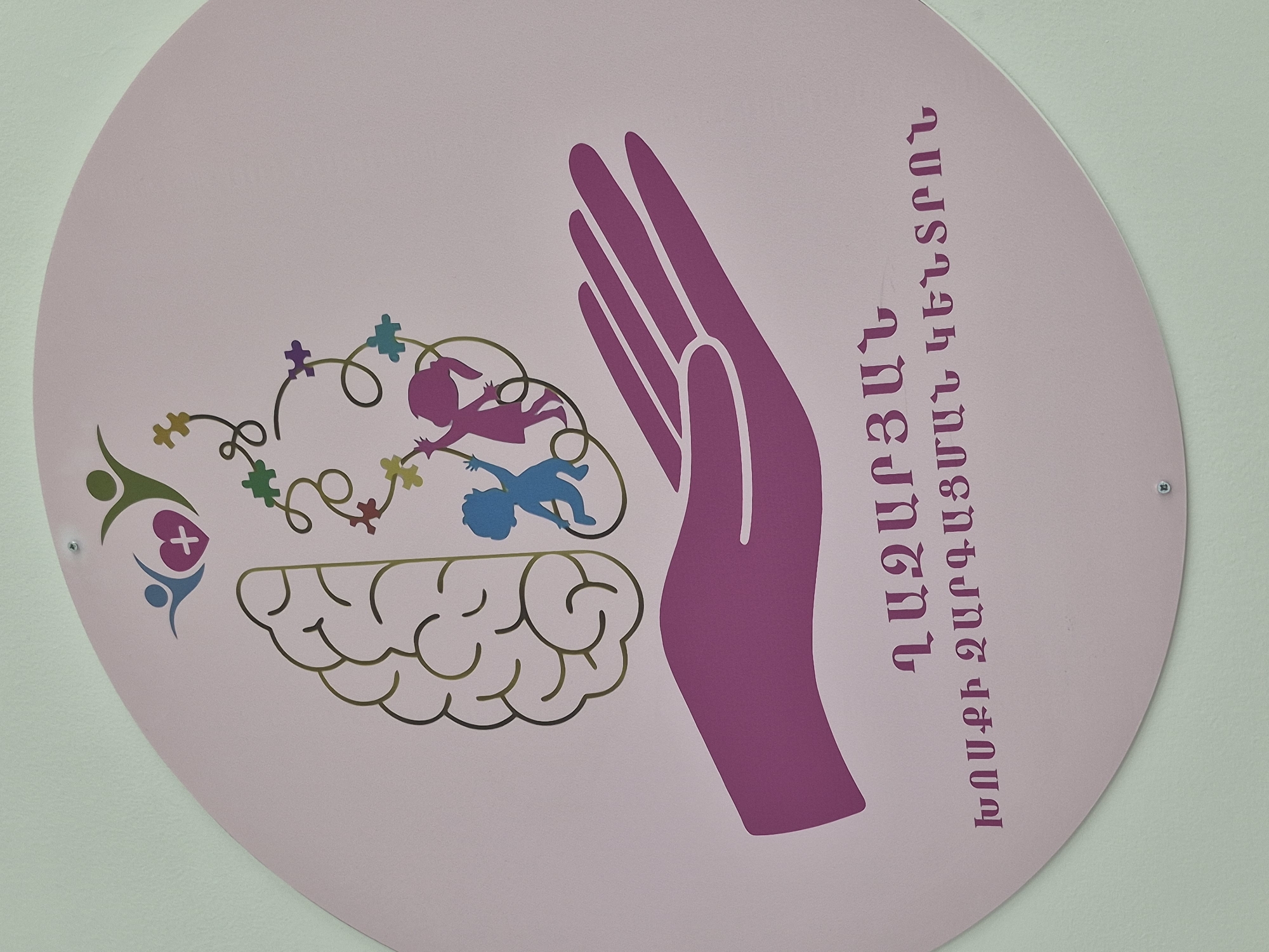 The image displays a large, light pink circular sign mounted on a light-colored wall, suggesting an indoor setting in Yerevan, Armenia.
The sign features a graphic design centered around themes of child development and support. A prominent purple human hand is depicted on the right side, fingers pointing upwards, symbolizing care and protection. Below and to the left of the hand, a brown outline of a human brain is shown. Above the brain, a swirling, thought cloud-like line is adorned with multiple colorful puzzle pieces, which are commonly associated with autism awareness. Inside this cloud, two stylized child figures, one magenta and one blue, are depicted, seemingly interacting with the puzzle pieces or the swirling lines, suggesting cognitive engagement and play.
On the far left, a smaller logo features two abstract human figures, one green and one blue, embracing a pink heart with a white cross inside, symbolizing community, health, and care.
Vertical text in Armenian script is present on the right side of the sign. It reads: "ՀԱԶԱՐԴԱՆԻ ԶԱՐԳԱՑՄԱՆ ԿԵՆՏՐՈՆ," which translates to "Hazaradan Development Center." The sign is fastened to the wall with two visible screws. The overall design and colors evoke a sense of warmth, inclusivity, and dedication to children's well-being and developmental support.