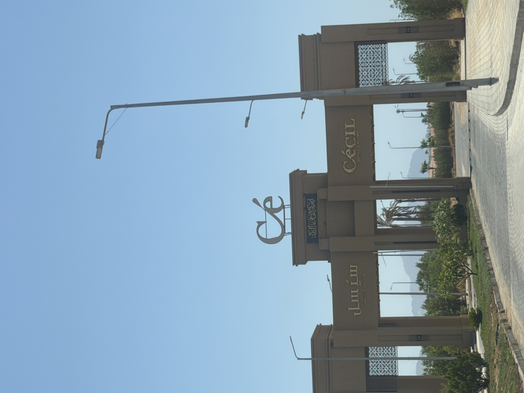 The image depicts the entrance to the Cecil Hotels & Resorts in Sharm El Sheikh, Egypt. The entrance is a large, ornate structure made of tan-colored stone, with two archways leading into the property. Above the archways, a prominent sign displays the hotel's name in both Arabic script and English. The Arabic script reads "سيسل," which translates to "Cecil." Above the English lettering "Cécil," there is a stylized logo that incorporates the letters "C" and "e."
The entrance is flanked by decorative metal gates and lush greenery, including trees and bushes. Beyond the entrance, palm trees and distant mountains are visible under a clear blue sky. Several streetlights are positioned along the road leading up to the entrance and within the resort grounds. The overall scene suggests a warm, sunny day in a tropical or desert environment. There are no people visible in the image. The perspective is from a vehicle approaching the entrance.