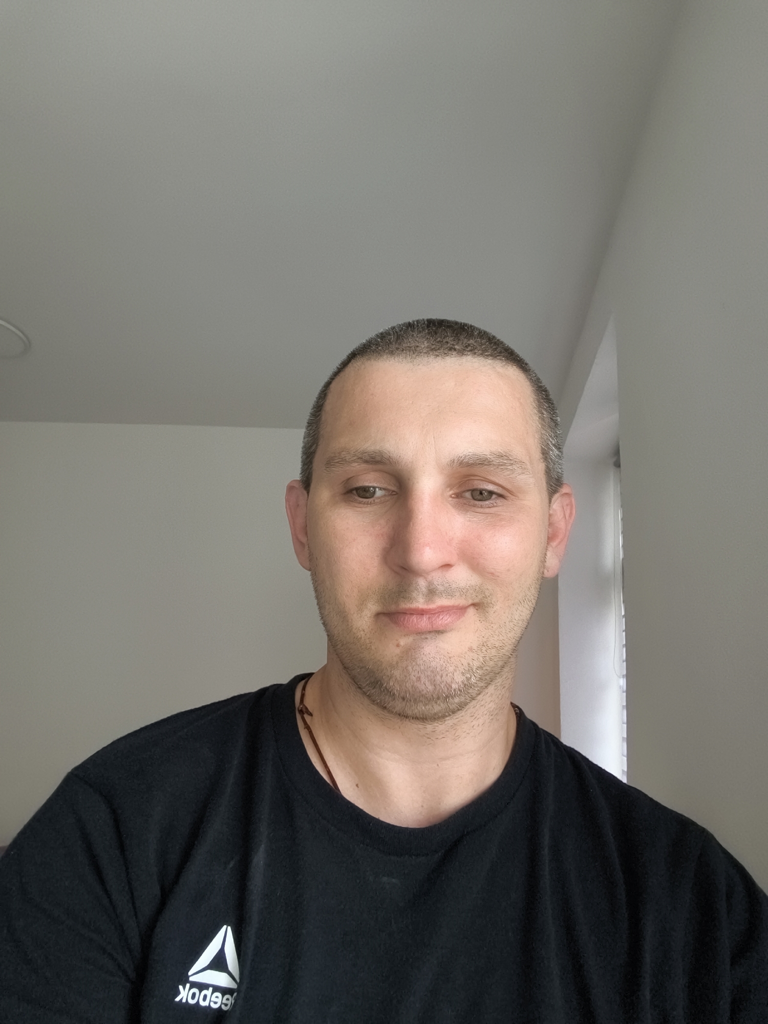 A close-up, head and shoulders selfie shows a white male with a buzz cut, light stubble, and light brown eyes. He is wearing a black t-shirt with a white Reebok logo on the left chest. He is looking down and to the right with a slight smile. The background is a plain, light-colored wall and ceiling, with a sliver of white vertical blinds visible on the right. The lighting is soft and even, suggesting an indoor setting, likely an apartment or house, as indicated by the presence of typical interior walls and ceiling finishes. The overall impression is of a casual, at-home portrait. The image does not provide any specific clues to its location within Wolgast, Germany, as it lacks any identifiable landmarks or distinguishing features of that city.