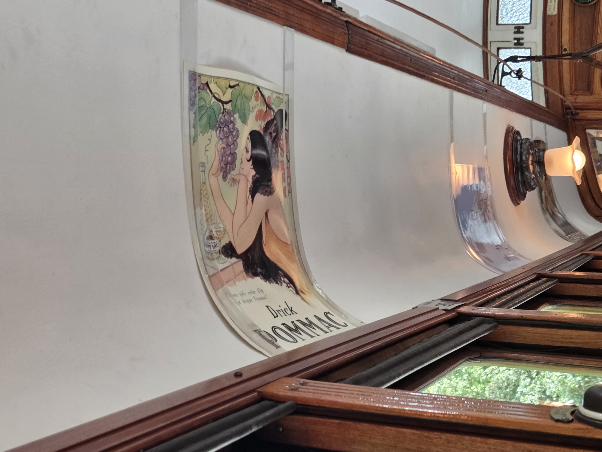 The image captures the interior of a vintage tram in Malmö, Sweden. The ceiling of the tram features a curved advertisement depicting a woman holding a bottle and grapes, with the text "Drink POMMAC" prominently displayed. The tram's interior is adorned with rich, polished wood paneling surrounding the windows and framing the ceiling advertisement. Outside the windows, lush green trees are visible, suggesting a pleasant outdoor environment. In the foreground, a ceiling light fixture with a decorative base illuminates the space. Further down the tram, two frosted windows with an "H H" motif are visible, along with a small sign indicating "stannar." The scene conveys a sense of nostalgia and historical charm.