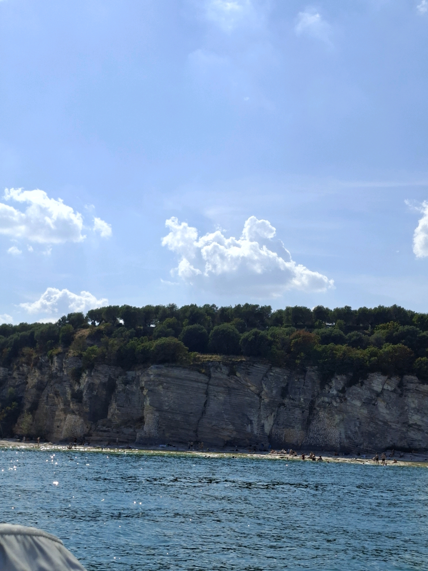 The image depicts a coastal scene with a prominent cliff face topped by dense green foliage. Below the cliff, a narrow beach is visible, populated by a number of individuals engaged in various beach activities, including sunbathing and swimming in the turquoise water. The sea is calm with a gentle ripple, and sunlight glints off its surface. The sky above is a clear blue, dotted with a few fluffy white clouds. The overall impression is one of a serene summer day at a popular seaside location. Given the distinctive rock formations and the lush vegetation, this scene is likely located on the southern shore of Lake Garda, possibly near the town of Sirmione, Italy, known for its dramatic cliffs and historical sites.