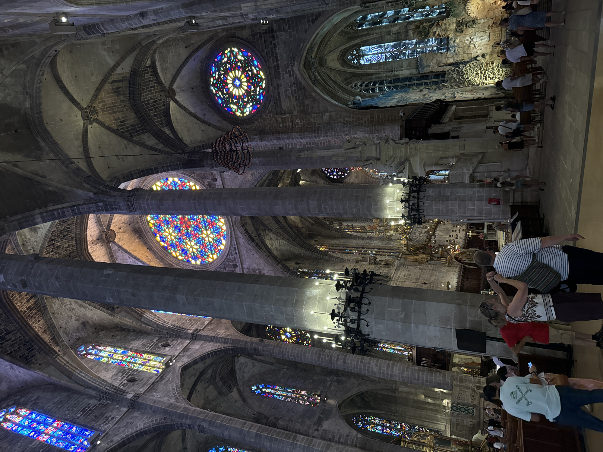 The photograph captures the interior of a grand Gothic cathedral, identified as the Cathedral of Santa Maria of Palma, located in Palma de Mallorca, Spain. The architecture is characterized by soaring stone columns, ribbed vaults, and numerous pointed arches, creating a sense of immense height and space.
Dominating the scene are several large, circular stained-glass windows, known as rose windows, and long, arched windows, all filled with vibrantly colored glass depicting intricate patterns and motifs. These windows cast colorful light throughout the cavernous interior.
Several large, stone pillars support the massive vaulted ceiling. Hanging from the ceiling are elaborate chandeliers and decorative light fixtures. In the foreground, a group of tourists are visible, some standing and observing, others engaged in conversation. One person in the foreground is wearing a light blue t-shirt with a graphic print and dark pants. Another person is wearing a striped t-shirt and carrying a bag over their shoulder. The floor appears to be paved with stone tiles.
The overall impression is one of awe-inspiring architecture and historical significance, with the interplay of light and shadow enhancing the dramatic atmosphere of the cathedral. The presence of tourists indicates that this is a public space and a popular tourist attraction.