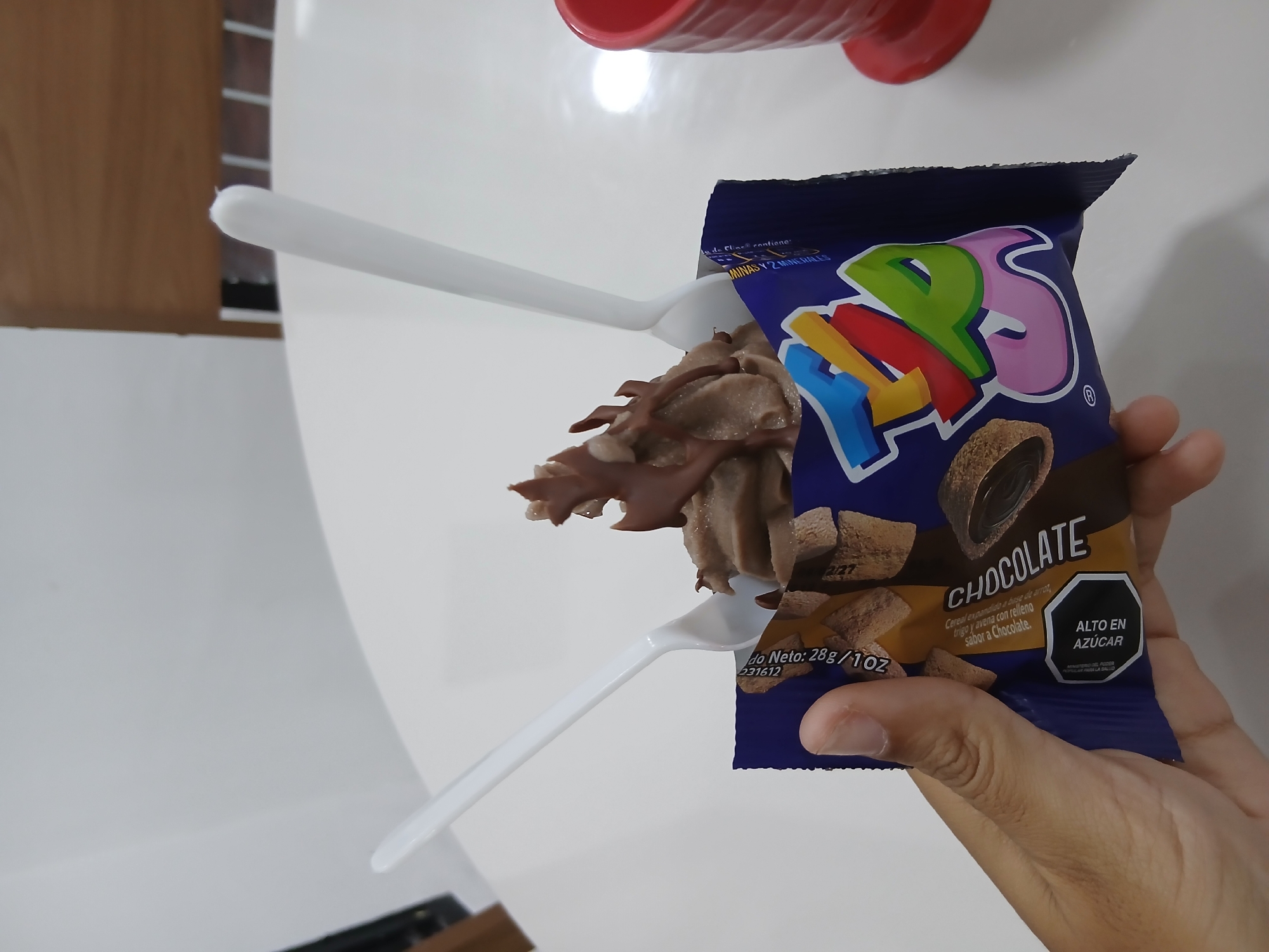A close-up, high-angle shot captures a human hand holding an opened snack bag of "FAPS" brand chocolate-flavored cereal. The dark blue bag, torn open at the top, is partially obscured by the hand and its contents. Chocolate-colored cereal pieces, described on the packaging as "Cereal expandido a base de arroz, trigo y avena con relleno sabor a Chocolate" (Expanded cereal made from rice, wheat, and oats with chocolate-flavored filling), are seen spilling out of the bag. Two white plastic spoons are positioned, suggesting the cereal is being eaten or prepared to be eaten directly from the bag.
The packaging clearly displays the brand name "FAPS" in colorful, bubbly letters. Below "CHOCOLATE", the net weight is listed as "28g / 1OZ". A prominent black octagonal warning label states "ALTO EN AZÚCAR" (High in Sugar), indicating a product regulated for its sugar content. Other visible text includes "MINASY 2 MINERALES" and what appears to be a batch or date code "231612" and "2/27".
The scene is set indoors, likely on a white table surface, with hints of a wooden surface and a red object (possibly a cup) in the background. The overall impression is of a casual snack moment in Juan Griego, Venezuela.