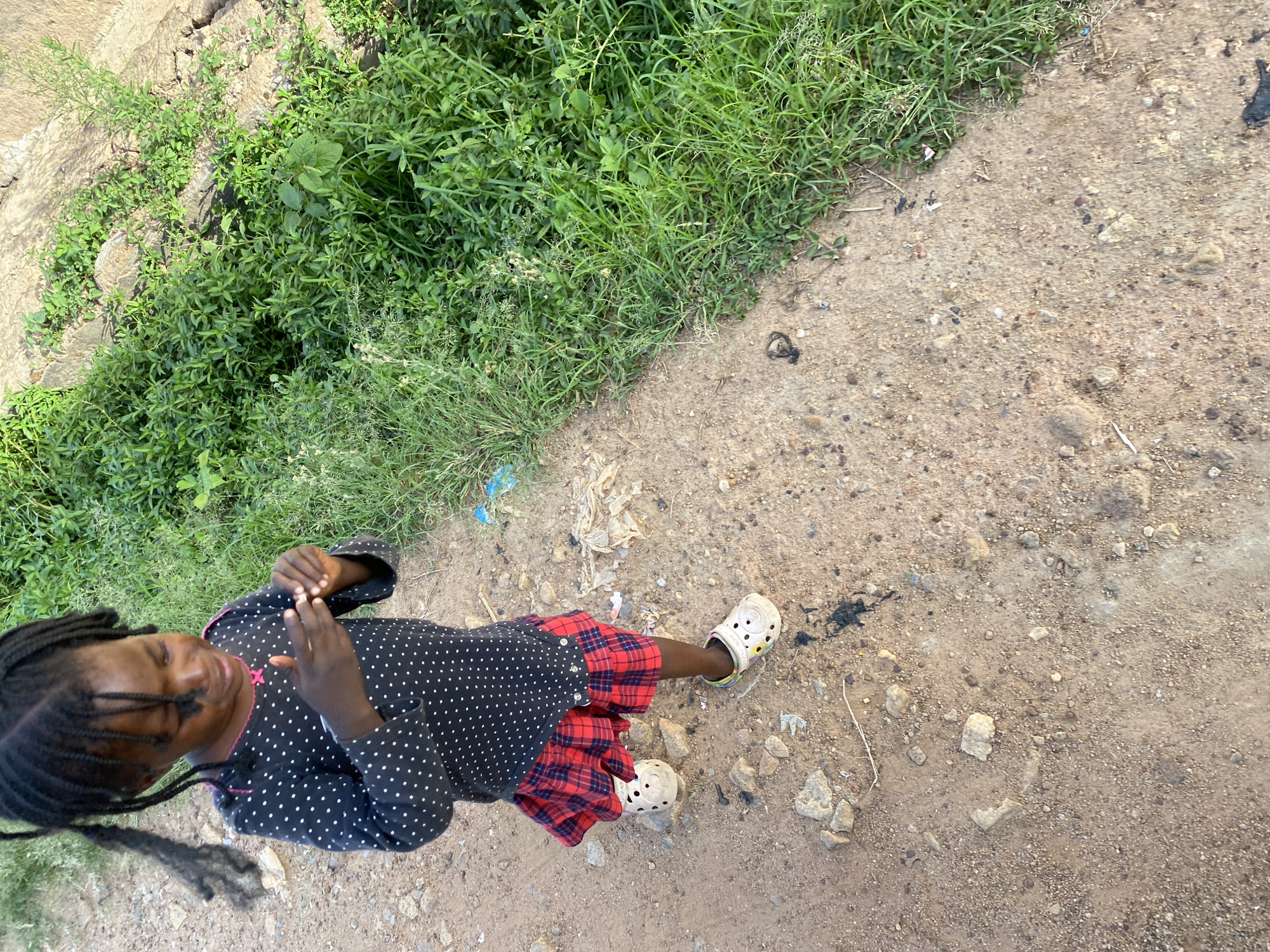The image depicts a young Black child in Bukuru, Nigeria, standing on a dirt and gravel path. The child wears a dark, polka-dot top, a red plaid skirt, and white Crocs. The path is bordered by overgrown green vegetation on the left. Debris and scattered rocks are visible on the ground.