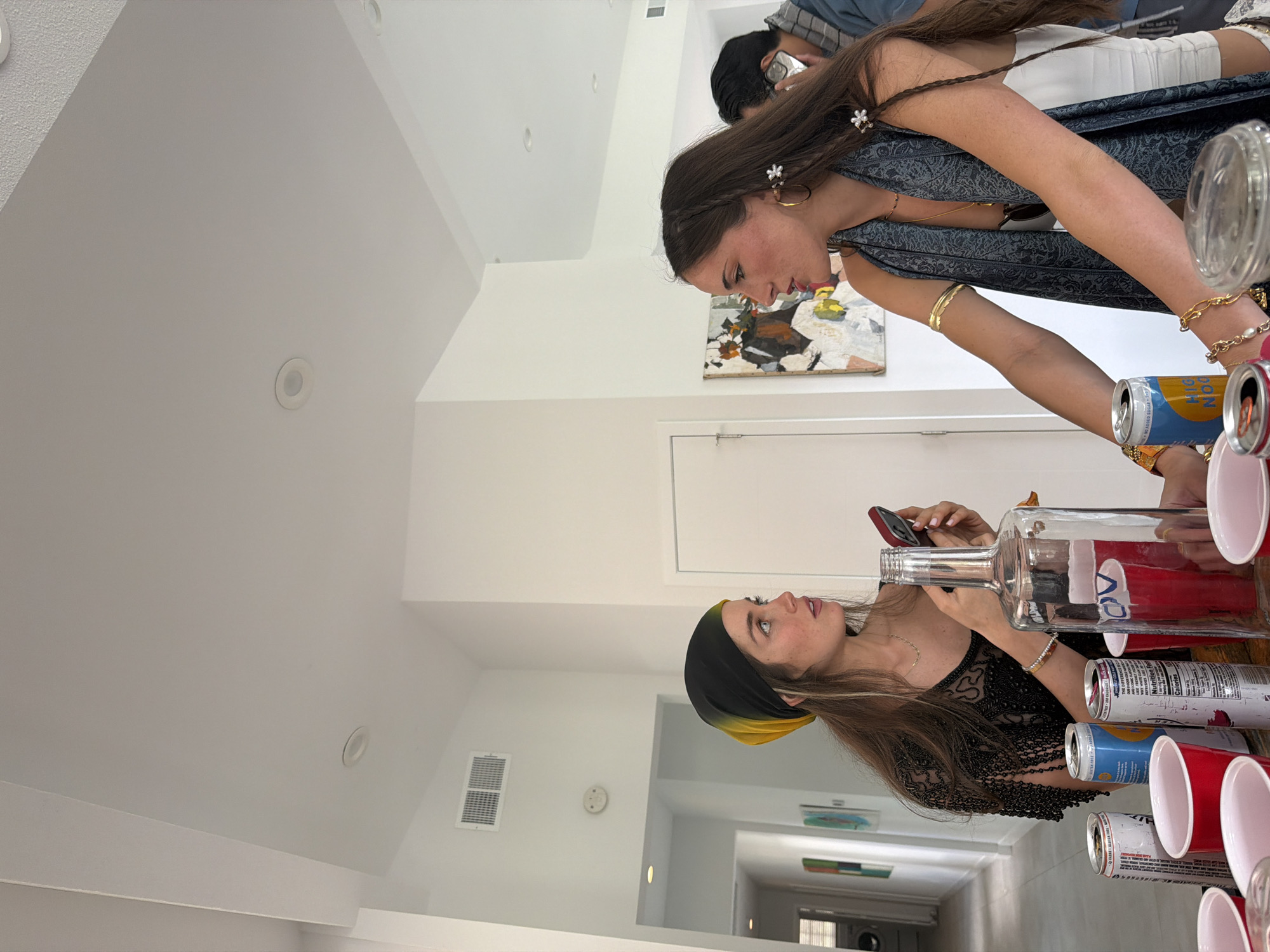 Two women are indoors in a well-lit room, likely at a party or social gathering. One woman is pouring a clear liquid from a large glass bottle into red plastic cups on a table. She is holding her cell phone with her other hand. She is wearing a black headscarf and a black, lacy top. The other woman is observing and helping. She is wearing a dark blue dress and multiple gold bracelets. There are cans of alcoholic beverages nearby, including High Noon. In the background is a white hallway with artwork on the walls and an open door leading to another room. The location is inferred to be in La Quinta, United States of America.