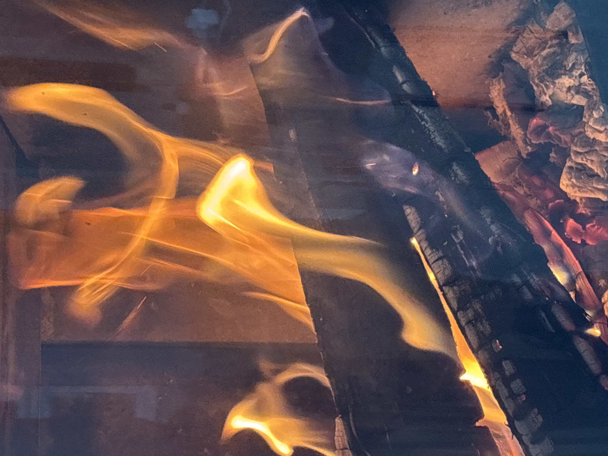 The image is a close-up of a fireplace fire, showing burning logs and flames. The flames are a vibrant orange and yellow, with some white hot areas. The logs are dark and charred, with glowing embers visible. The background is dark and out of focus, suggesting a dimly lit room. There are no people visible in the image. The scene is indoors, within a fireplace. There is no information in the image to suggest a specific location in Zvenigorod, Russia.