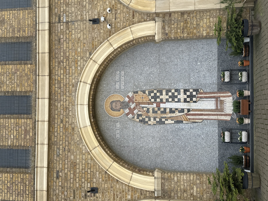 The image displays the exterior facade of the Saint Sava Serbian Orthodox Church, located on Lancaster Road in London, United Kingdom. Central to the scene is a large, full-body mosaic of Saint Sava, rendered in tesserae of various colors, depicting a bearded figure with a golden halo, dressed in ornate ecclesiastical robes, holding a closed book in his left hand. Cyrillic text above the figure identifies him as "Sveti Sava" and "Prvi Srpski Arhiepiskop" (First Serbian Archbishop). This mosaic is set within a prominent arched recess on a light-colored brick wall. Above the arch, three rectangular windows with dark grilles are visible, beneath which anti-bird spikes are installed. The ground directly in front of the facade is paved with interlocking brick pavers. Symmetrically arranged along the base of the mosaic are various potted plants and flowers, some placed on tiered black metal stands and others in individual pots or a rectangular wooden planter. Two larger potted evergreen trees in dark wooden barrels flank the entire arrangement. On the right side of the arch, a black mounting bracket holds two white CCTV cameras directed downwards. A black wall-mounted light fixture is visible on the left side, outside the arch.