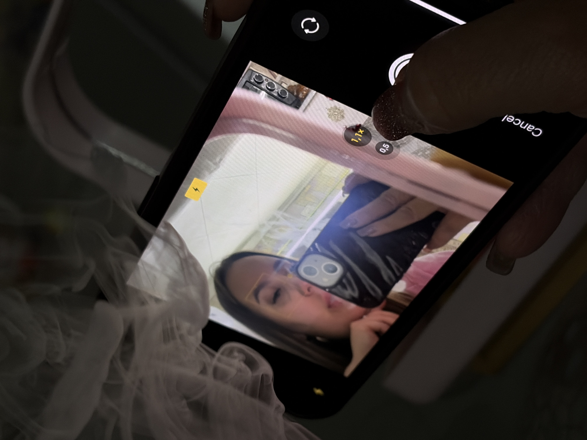 The image shows a black smartphone held by a person with nail polish, displaying a selfie being taken in a bathroom or similar space. The selfie captures a woman holding another smartphone, her face reflected in a mirror. A translucent, wispy element, possibly smoke or mist, appears near the bottom left of the image. The background as seen in the mirror reflection appears to be a domestic kitchen setting. No evidence exists within this image to pinpoint the location within Port-Katon, Russia.