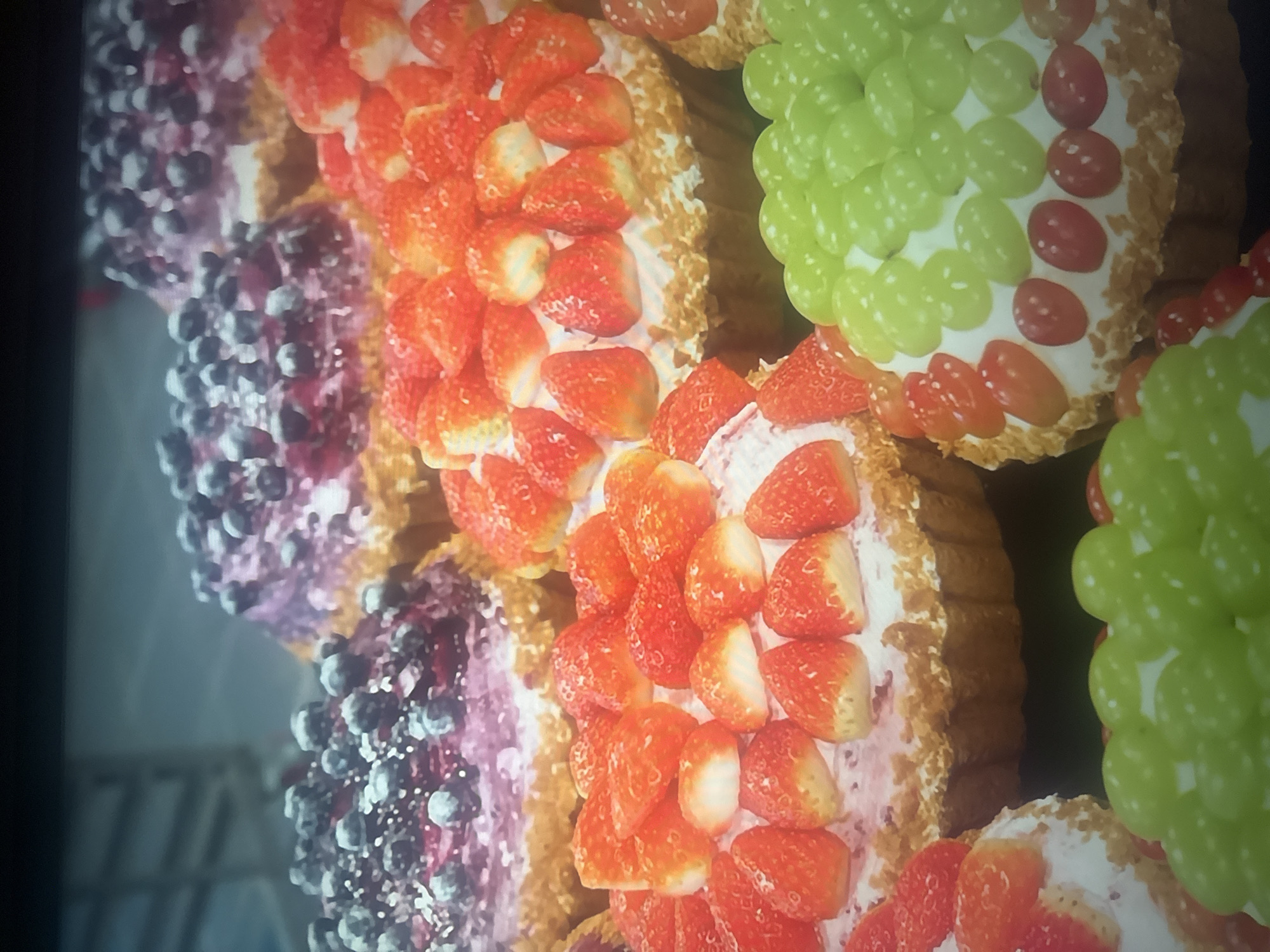 In Herzliya, Israel, a close-up shot captures a display of artisanal fruit tarts. The tarts feature various toppings, including fresh strawberries, blueberries, and green grapes, arranged atop creamy fillings within golden crusts. Some tarts are also adorned with smaller red grapes, creating a vibrant mosaic of colors and textures. The background is blurred, suggesting the tarts are likely displayed in a bakery or market setting, though no specific location within Herzliya is identifiable. There are no people or animals visible in the image.