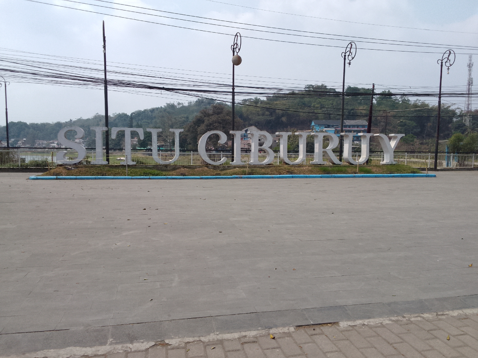 A wide-angle, eye-level outdoor shot captures the "SITU CIBURUY" signage in Padalarang, Indonesia. The signage is composed of large, white, three-dimensional letters mounted on a grassy area, with a low blue and white barrier in front. Behind the sign, a body of water is visible, reflecting the overcast sky. Trees and buildings dot the landscape in the background, with hills rising behind them. Overhead, a dense network of electrical wires crisscrosses the frame, supported by utility poles. Decorative lampposts with globe-shaped lights stand near the signage. The foreground consists of a paved area, likely a pedestrian walkway or plaza, made of grey square tiles. The overall scene is bright, suggesting daytime.