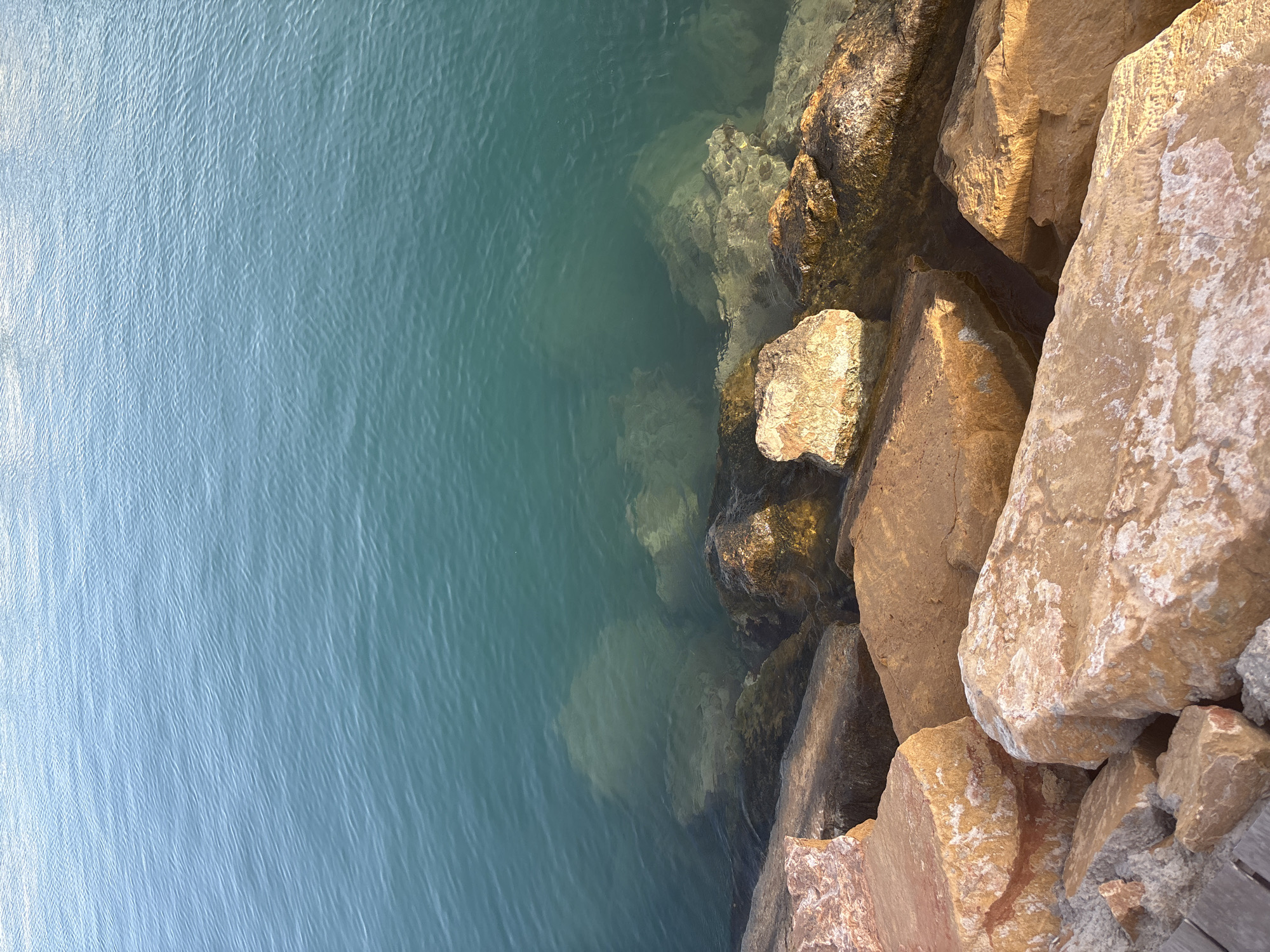 A high-angle, close-up shot captures a section of a body of water meeting a rocky shoreline. The water, a vibrant teal-blue, has a rippled surface, indicating light movement. Submerged rocks are visible beneath the clear surface, adding depth and texture. The shoreline is composed of large, irregularly shaped rocks in shades of tan, orange, and brown, with some lighter mineral deposits. A small portion of what appears to be a wooden plank walkway is visible in the lower left corner, peeking out from beneath the rocks. The scene is well-lit, suggesting daytime. The location is Unknown, Unknown. There are no people or animals visible.