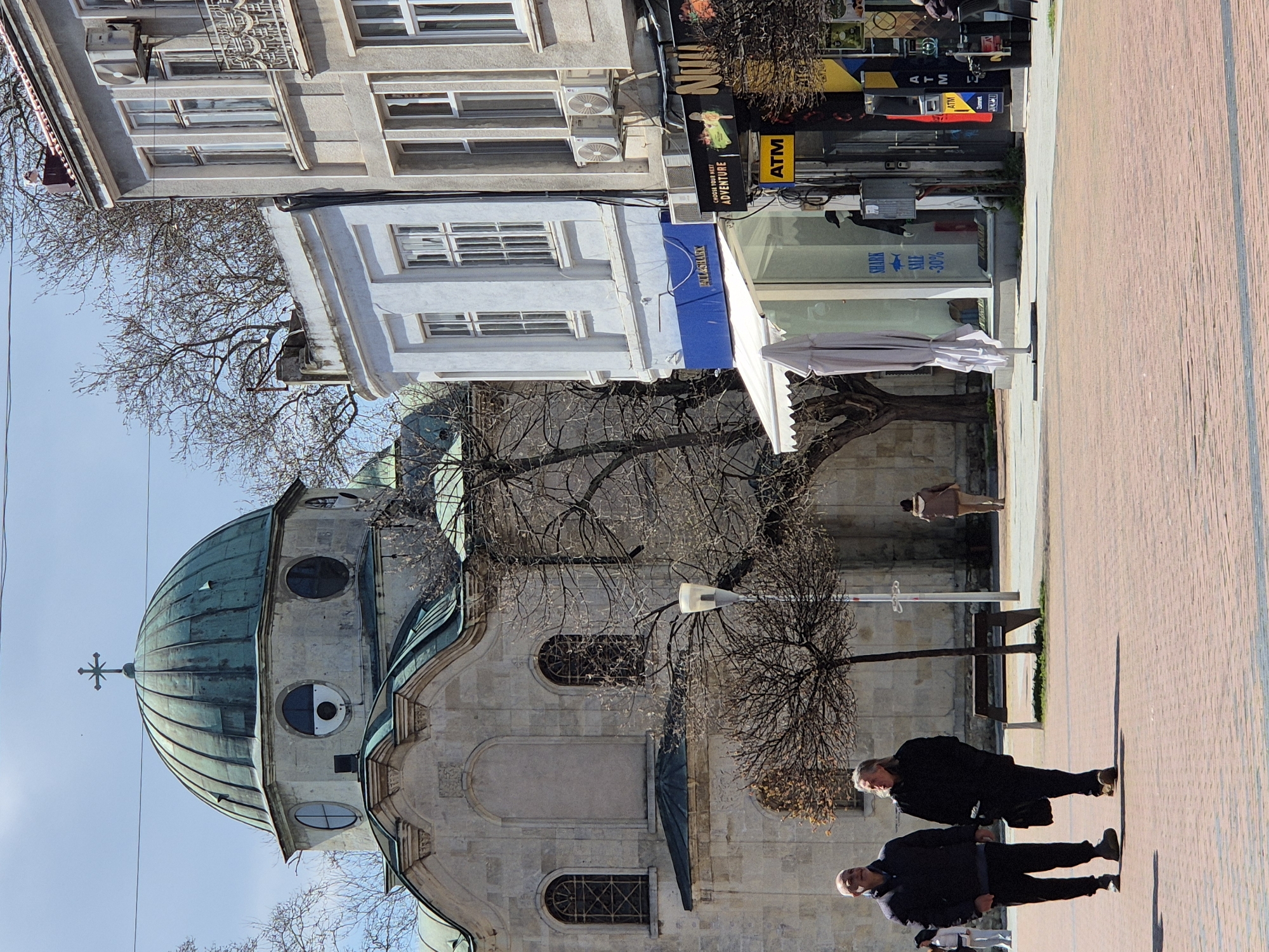 The image shows a street scene in Varna, Bulgaria. A green-domed church stands prominently on the left. Two men are walking along the brick-paved sidewalk on the right. A shop with an "ATM" sign and a blue awning is visible behind them. The trees are bare, suggesting it is either late autumn or winter. Buildings are on the far side of the church. There is also a street lamp and a park bench. Another person can be seen walking in the distance.