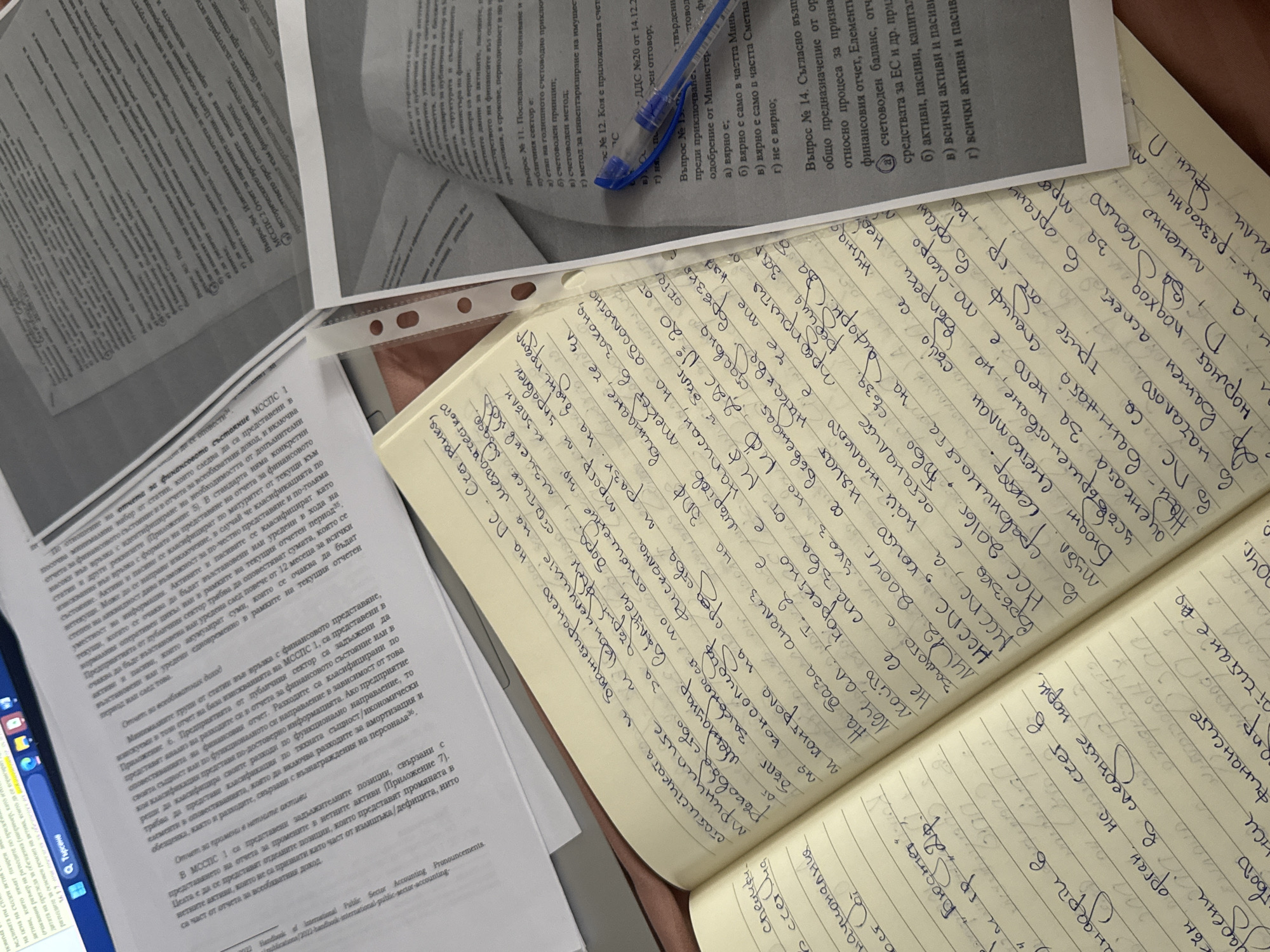 The image captures an overhead view of a study or work desk, featuring several printed documents, an open notebook, and a blue ballpoint pen. The setting appears to be indoors, well-lit, suggesting daytime.
The main subjects are various papers and a notebook, indicating an activity involving reading, learning, or working. One document is folded and partially obscures another, while a third is open and covered with handwritten notes in a spiral-bound notebook. A blue pen rests on one of the printed pages.
The printed documents contain text primarily in Bulgarian Cyrillic script, with titles and questions related to accounting, finance, and the public sector. Visible text includes phrases like "Въпрос №3. При подготовката на финансовите отчети..." (Question #3. When preparing financial statements...), "МССПС 2 Отчет за паричните потоци" (IPSAS 2 Statement of Cash Flows), "Въпрос № 11. Последваното оценяване и публичния сектор е:" (Question #11. Subsequent measurement and public sector is:), and mentions of "финансовото състояние" (financial position), "публичния сектор" (public sector), "активи и пасиви" (assets and liabilities), and "бюджет" (budget). One document explicitly references "Handbook of International Public Sector Accounting Pronouncements."
The handwritten notes in the notebook are also in Bulgarian Cyrillic, discussing topics such as "Нормативен подход" (Normative approach), "Бюджетирането на ПС" (Budgeting of the Public Sector), "финансите" (finance), "счетоводния" (accounting), "статистиката" (statistics), and references to "МССПС" (IPSAS) and "НСС" (National Accounting Standards). A partially visible hand, possibly belonging to the note-taker, is seen holding the notebook.
The overall scene depicts an academic or professional environment focused on financial or public sector accounting studies in Mechkarevo, Bulgaria.