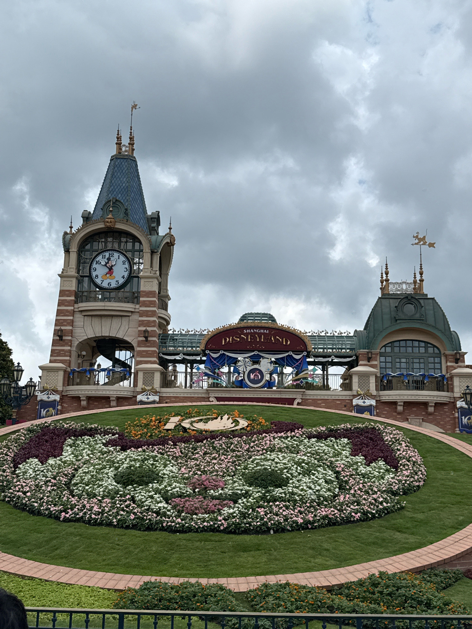 The image shows the entrance to Shanghai Disneyland in Pudong, China, under a cloudy sky. In the foreground, a large circular flower bed is arranged in the shape of Mickey Mouse's head, composed of white, pink, and dark purple flowers, with a dark green hedge forming the outline. Behind the flower bed is a paved pathway. A brick structure with arched openings and decorative banners stands on either side. Prominently featured is a tall clock tower with a large clock face displaying Mickey Mouse. Above the entrance to the park, a sign reads "SHANGHAI DISNEYLAND" in a curved, ornate font, adorned with banners and the "10" logo for the park's 10th anniversary. The background features more of the park's architecture, including arched windows and a green domed roof. The overall scene depicts the festive and iconic entrance to the theme park.