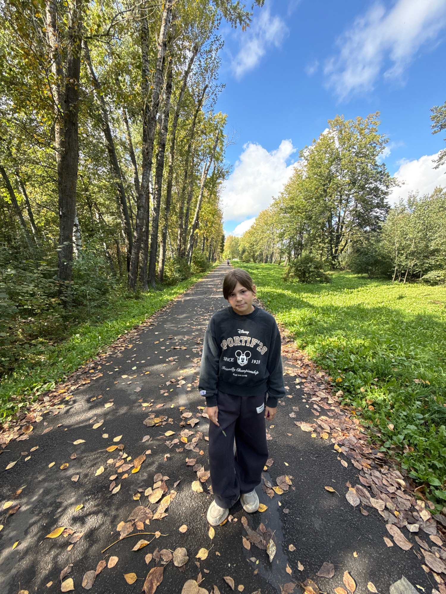 A young person stands on an asphalt path in Rusanovo, Russia, during autumn. The path is lined with tall trees with green and yellowing leaves, suggesting the fall season. Fallen leaves are scattered across the path and the surrounding grassy area. The sky is blue with white clouds. The person is wearing a dark sweatshirt with a graphic print and dark pants, along with light-colored sneakers. The person is facing the camera and looking directly at it. The overall lighting indicates it is daytime.