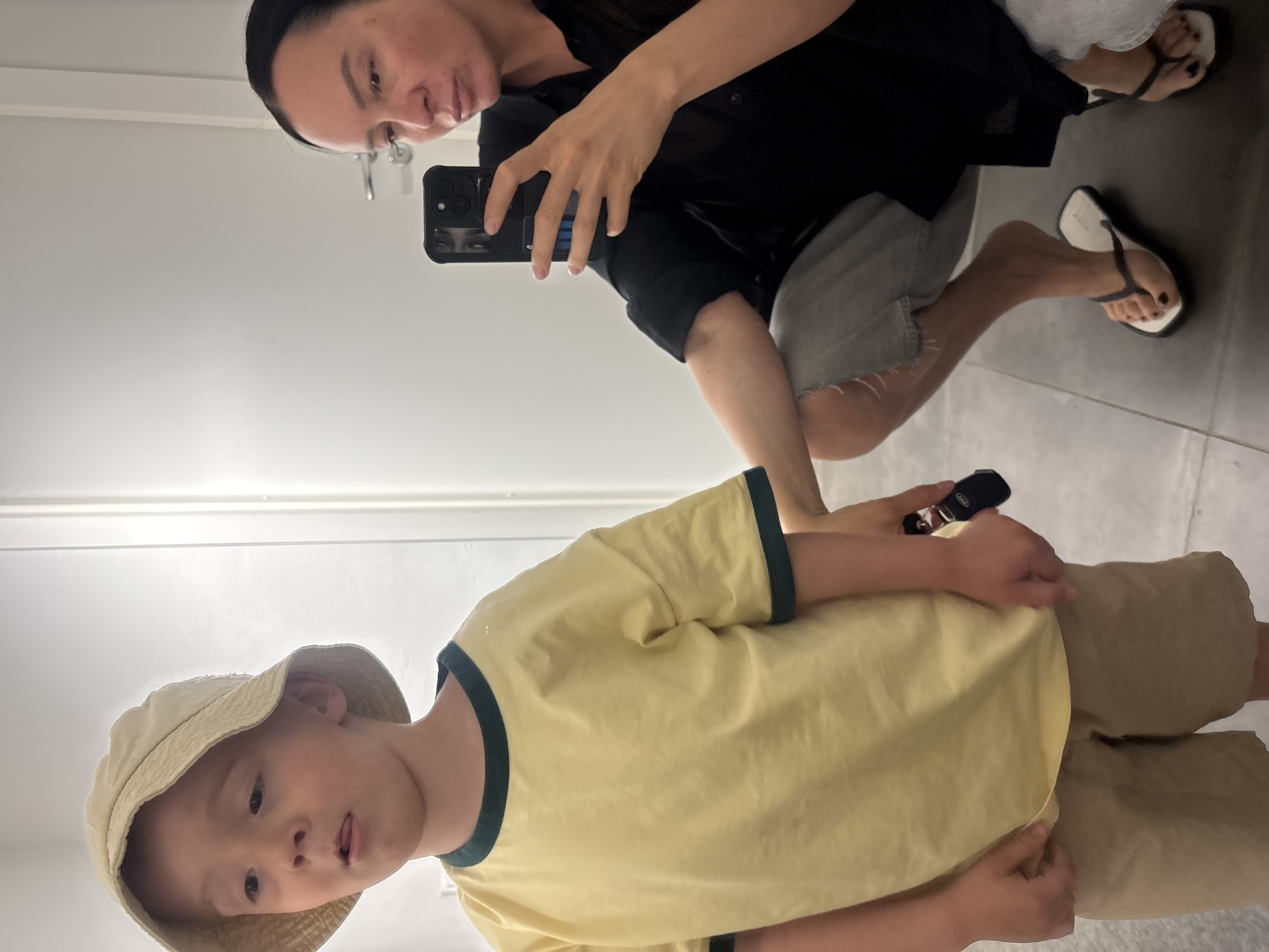 A woman is holding a smartphone and taking a picture of a child. The woman is wearing a black shirt and gray shorts with flip-flops. The child is wearing a beige bucket hat, a yellow t-shirt with dark green trim, and beige shorts. They are in an indoor space with white walls and light gray tiled floors. The location in the city of Chaikovsky, Russia, cannot be determined from the given visual information.