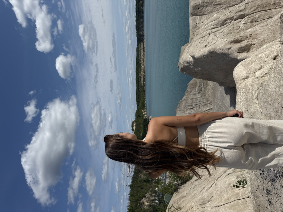 A female individual is positioned with her back to the viewer, standing on pale, eroded terrain overlooking a large turquoise-colored body of water under a bright blue sky with scattered white cumulus clouds. The scene is located at Druzhba Lake, a flooded former quarry near Druzhba, Ukraine. The individual has long, dark brown, wind-blown hair cascading down her back and wears a white halter-neck crop top and light beige, loose-fitting trousers. She is gazing across the water towards the distant forested banks that encircle the lake. The immediate foreground consists of rugged, light-colored, sculpted earth formations, characteristic of a quarry environment.