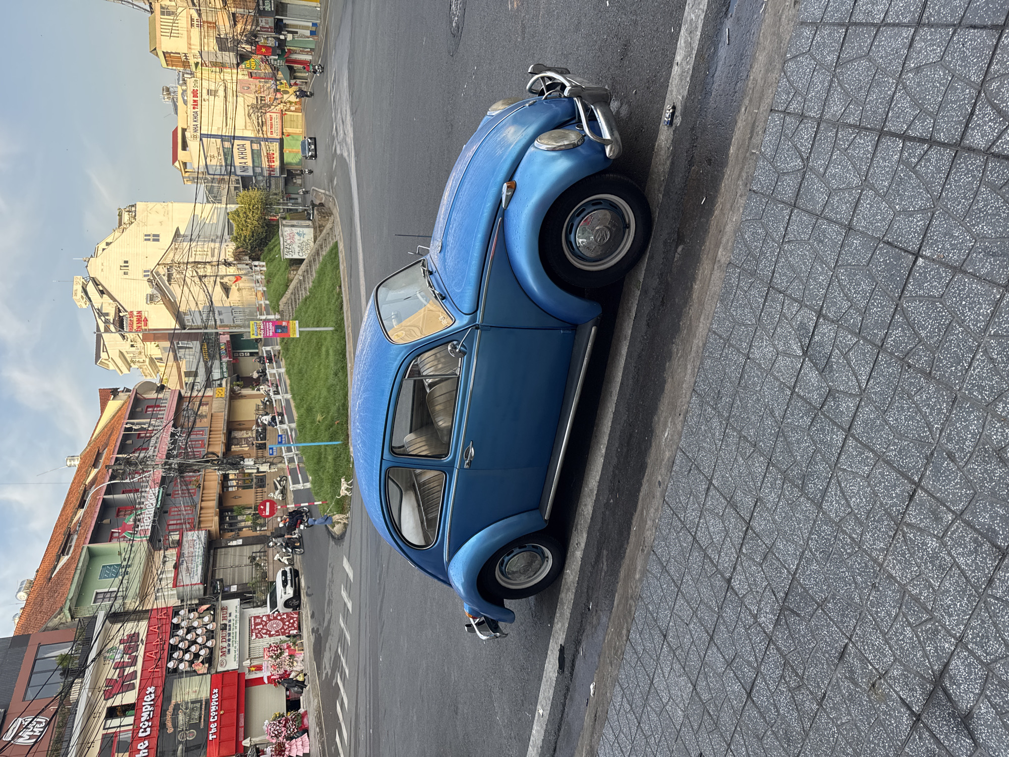 This vibrant image captures a bustling street scene in Da Lat, Vietnam, under clear daylight conditions. The main subject is a classic, light blue Volkswagen Beetle, parked parallel to a textured gray sidewalk. The car appears to have water droplets or dew on its surface, featuring chrome bumpers, headlights, and hubcaps.
The street is lined with multi-story buildings that exhibit varied architectural styles, many with balconies and colorful facades. Numerous electrical wires crisscross overhead, a common sight in Vietnamese cities. On the sidewalks, multiple motorcycles are parked, and others are visible moving on the asphalt road. A grassy median or embankment runs along one side of the street, where a person is seen walking a small, light-colored dog on a leash. Another small dog is visible farther down the grassy patch.
The scene depicts everyday urban activity, with businesses operating and people and vehicles moving through the city. The time of day appears to be morning or early afternoon, given the bright, clear sky.
Visible text on storefronts and signs includes several instances of "NHA KHOA" (Dentist), with one specifically reading "NHA KHOA TÂM ĐỨC Smile." Other signs display "The Complex" multiple times, "MỸ DŨNG HUẾ," and "AN NHÂN." A price of "199.000" (likely Vietnamese Dong) is visible on a yellow sign. The overall atmosphere suggests a lively commercial district.