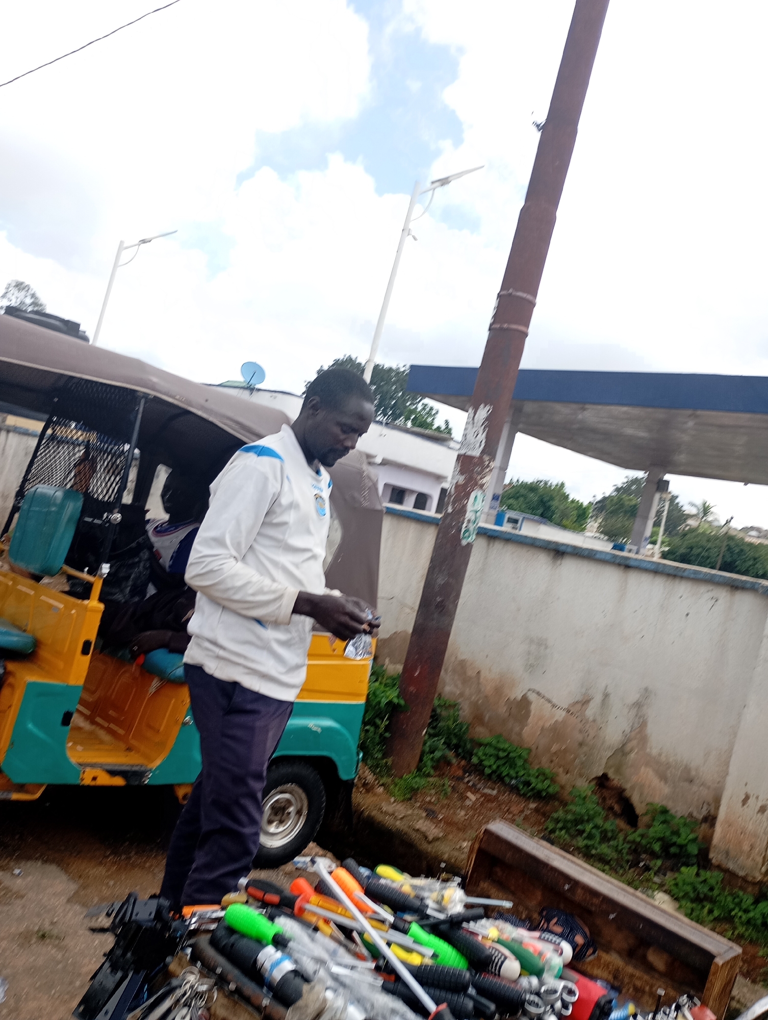 In Jos, Nigeria, a man in a white and blue jersey and dark trousers stands near a yellow and teal auto-rickshaw. He is holding a small object in his hands and appears to be inspecting it. The auto-rickshaw has its door open, revealing a dark interior with some blue cushioning. In the foreground, a collection of tools, including screwdrivers with colored handles, are scattered on the ground. Behind the man and the auto-rickshaw, a textured wall stretches horizontally. A tall, reddish-brown pole stands prominently on the right side of the image, leaning slightly. In the background, a building with a dark blue overhang and white walls is visible, with green foliage and trees surrounding it. The sky above is a mix of white clouds and blue sky.