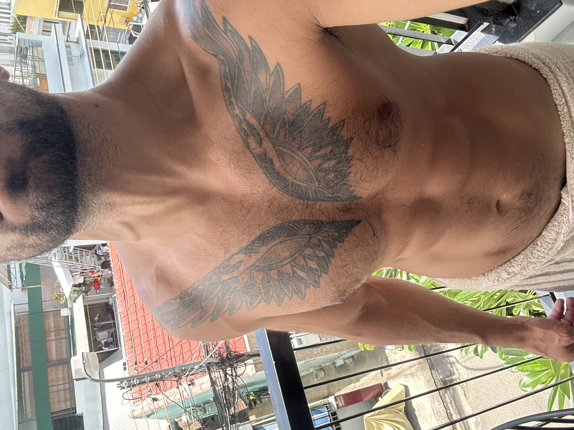 A man with a beard and an angel wing tattoo on his chest stands on a balcony overlooking a city in Nha Trang Ward, Vietnam. The man is wearing a light-colored towel around his waist. The buildings in the background are colorful and have red tiled roofs. There are power lines visible, indicating an urban environment. The balcony has a black railing and the man is standing in sunlight.