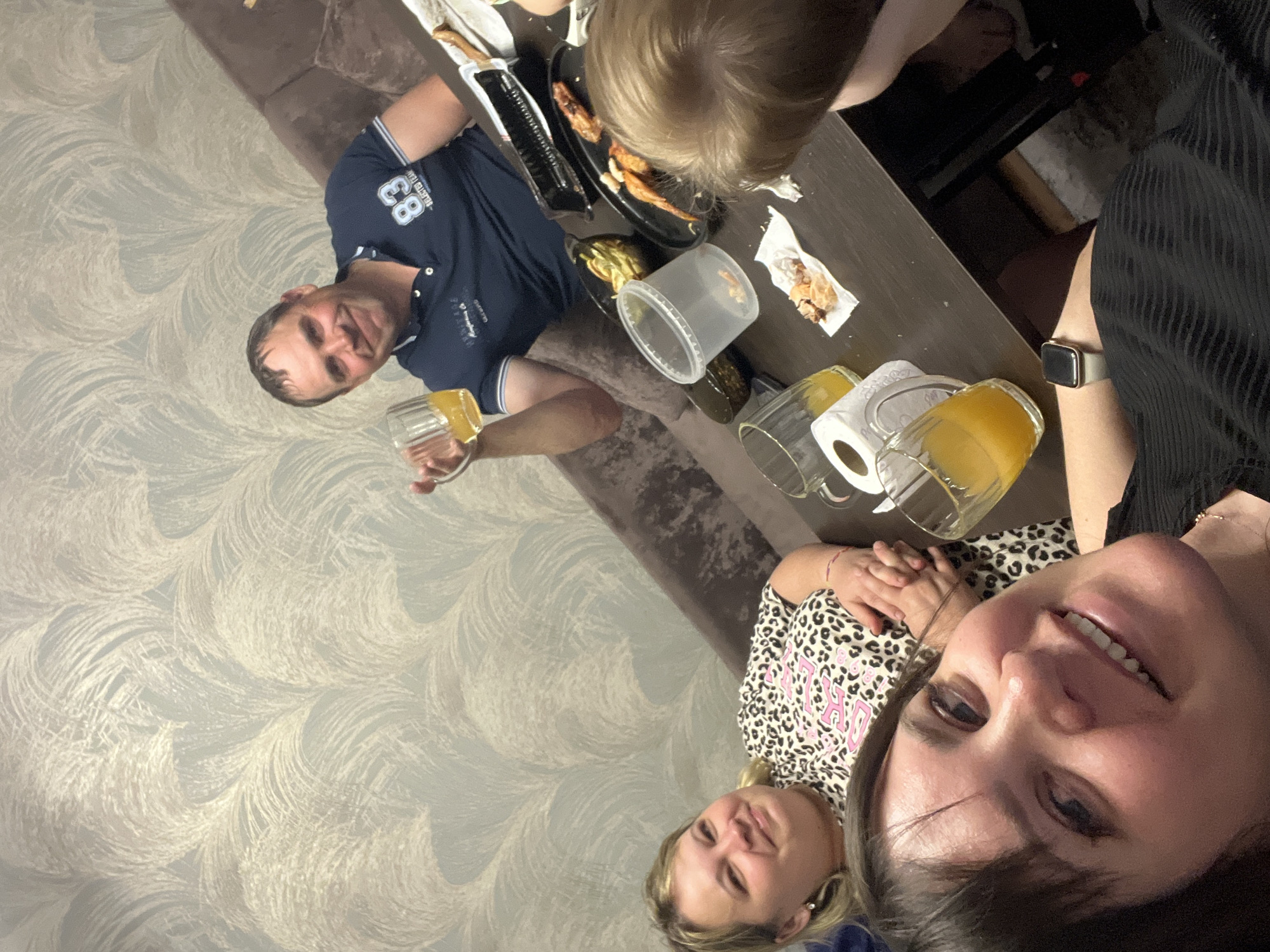 A family is having a meal together indoors. There are at least four individuals visible: two women, one man, and a child. The man, positioned on the left side of the image, is holding a glass of yellow liquid. He is wearing a blue polo shirt with the number "83" on it. The women are located on the right side of the image, one of whom is taking the picture. She is wearing a black outfit and a wristwatch. The other woman is wearing a leopard-print shirt. The child is seated at the top of the frame.
A table in the center of the image is holding a plate of food, a plastic container, and several glasses with a yellow liquid. There is also a roll of paper towels on the table.
The location is implied to be indoors, possibly a home, given the presence of a sofa and patterned wallpaper. There is no other information in the image to confirm that it is located in the city of Noviy, Russia.