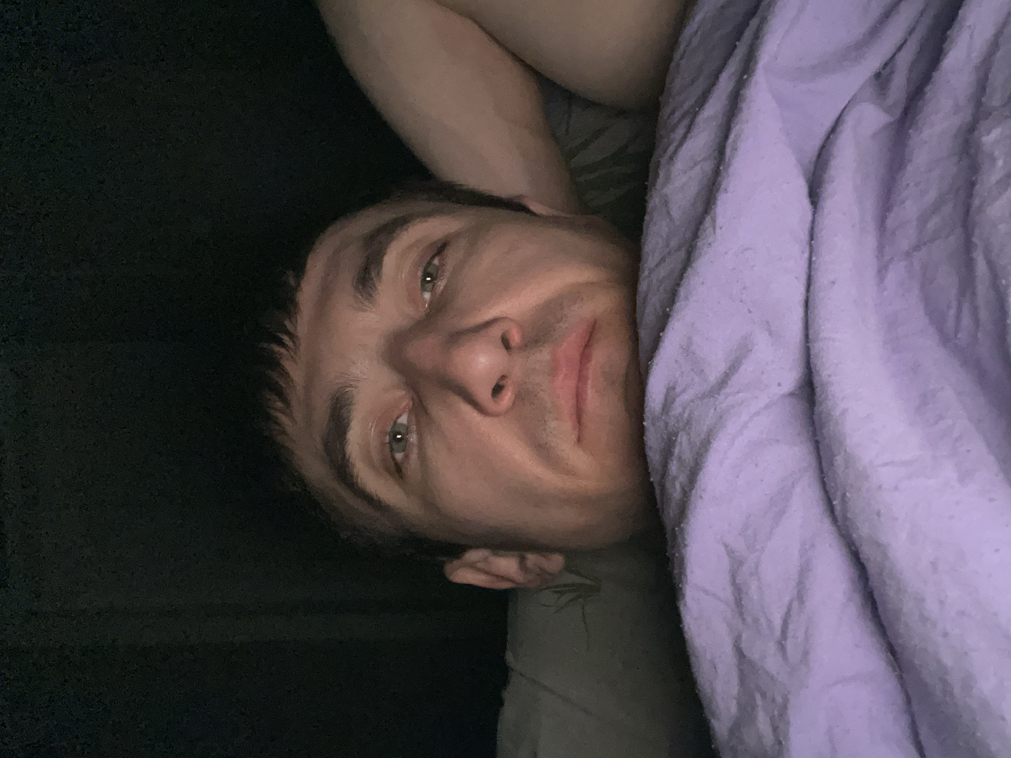 A man is lying in bed under a purple blanket, looking at the camera. His arm is bent behind his head, and he is resting his head on his arm. The background is dark, suggesting it may be night or that the room is dimly lit. The man has short brown hair and green eyes. The image was taken indoors in a bedroom.