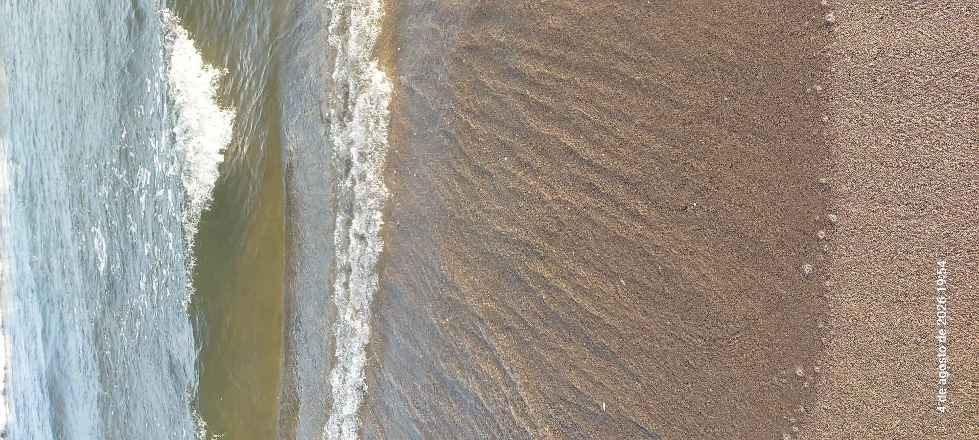 The image shows a close-up view of a sandy beach at Castelldefels, Spain, with gentle waves lapping at the shore. The sand is wet and textured, showing ripples and patterns left by the receding water. Small white bubbles are visible at the edge of the water. The water itself is a murky green-brown color, with a white foam line marking the crest of a breaking wave. The sky is not visible. A timestamp in the bottom left corner reads "4 de agosto de 2026 19:54," indicating the photo was taken on August 4, 2026, at 7:54 PM. There are no people or animals visible in the frame.
