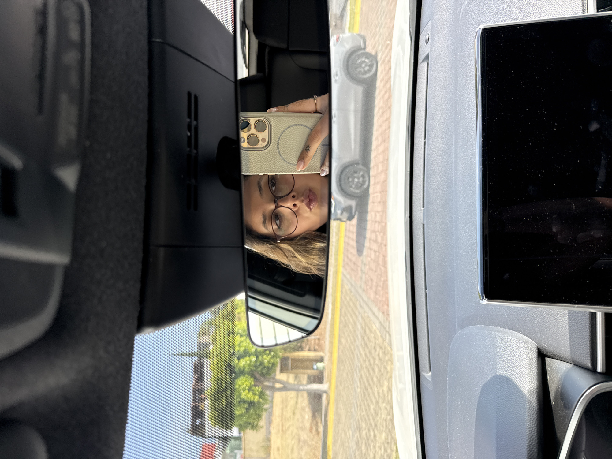 A selfie is reflected in a rearview mirror inside a vehicle, presumably a car. The selfie shows a person with brown hair, glasses, and a phone with a tan case, making a kissing face. The rearview mirror also reflects an outside environment showing a street with trees and buildings. Based on visual markers of architecture and vegetation, it is likely that this image was taken in the city of Puebla, Mexico. A toy car and an electronic device with a black screen are positioned in the foreground.