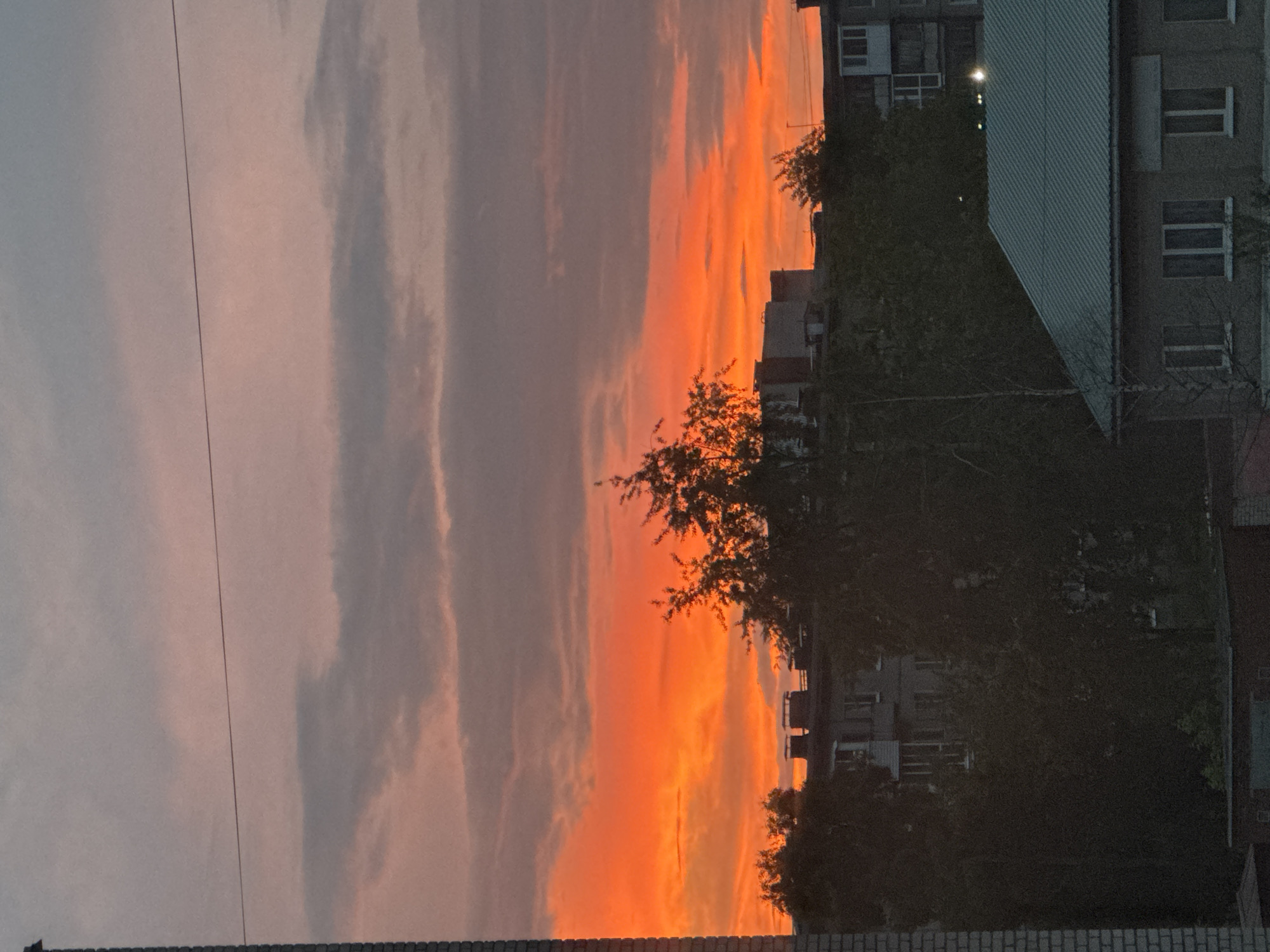 The image depicts a vibrant sunset over a city skyline, likely Solikamsk, Russia, given the context. The sky is a dramatic gradient of orange and pink hues, with streaks of gray clouds scattered throughout. Silhouetted against this fiery backdrop are the rooftops and upper floors of apartment buildings and other structures. Dense foliage of trees partially obscures some of the buildings, creating a contrast between the natural and urban elements. The foreground features a portion of a brick structure on the left, possibly a building or chimney, and a dark, lower-lying building with a corrugated metal roof on the right. The overall impression is a serene, yet striking, end-of-day scene in an urban environment.