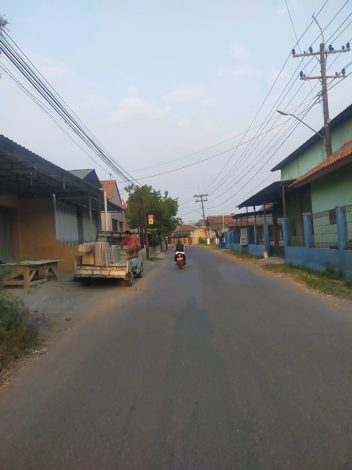 In Purbasana, Indonesia, a truck loaded with boxes is parked on the side of a paved road. A person wearing a red shirt is sitting on the edge of the truck bed, tending to the boxes. Further down the road, a motorcycle with a rider is visible. The scene is lined with houses and utility poles, with trees and vegetation present along the roadside. The sky is clear and bright, suggesting daylight.