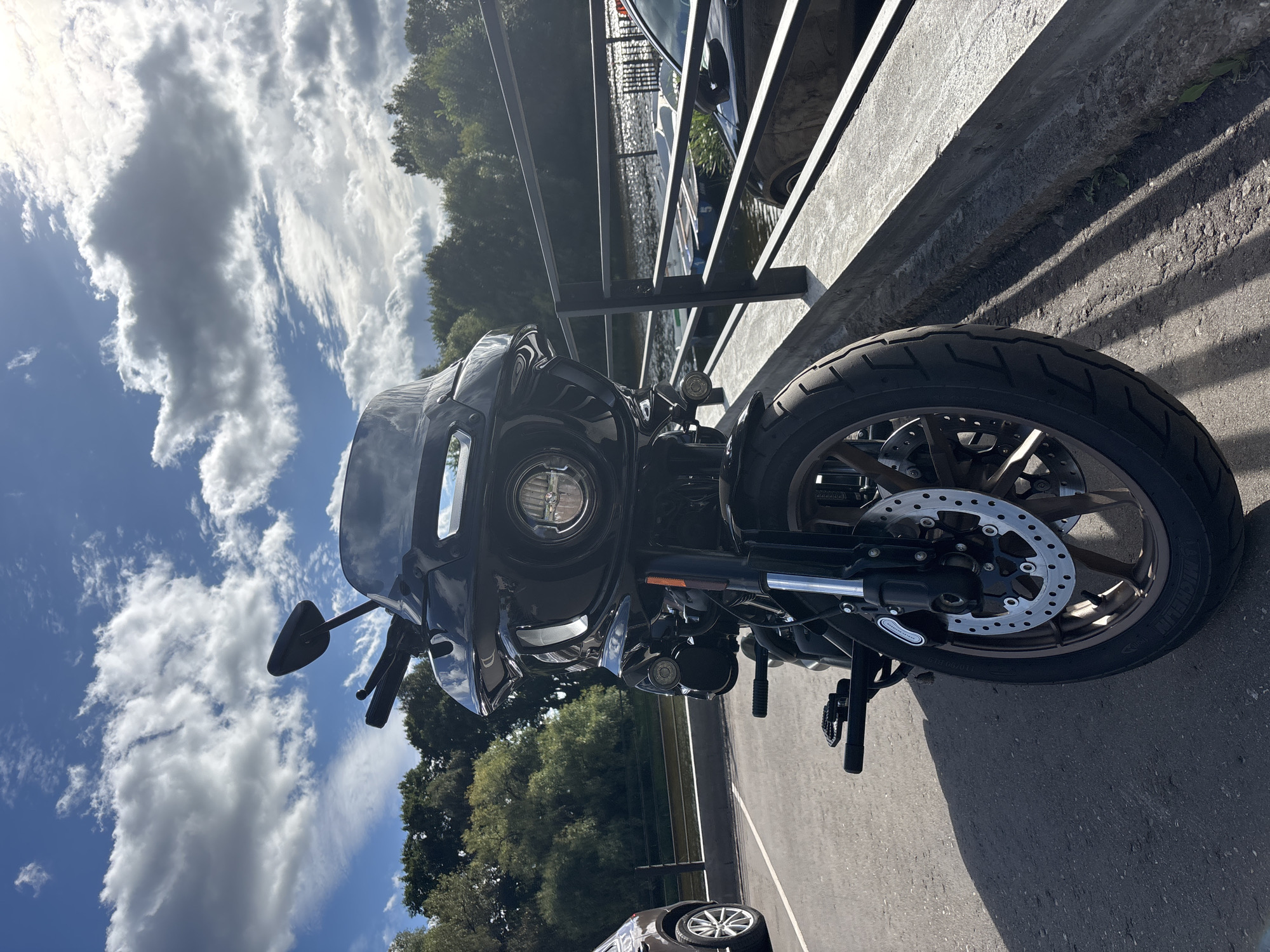 A black motorcycle is parked on an asphalt surface next to a concrete embankment with a metal railing. Behind the railing is a body of water with several boats on it, and beyond that, a line of trees. The sky above is blue with scattered white clouds. The motorcycle is positioned facing forward, slightly angled to the right. A dark-colored car is visible to the left of the motorcycle. The scene appears to be a sunny day, possibly in a park or recreational area. The location is identified as Zvenigorod, Russia.