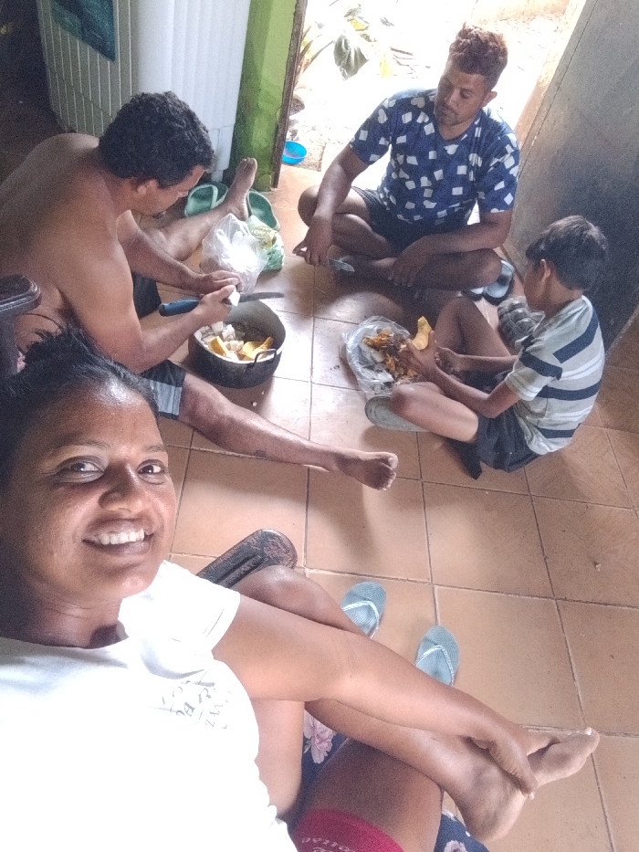 Four people sit on a tile floor in a room. A smiling woman in a white shirt takes a selfie, her legs crossed. To her right, a shirtless man peels food into a metal pot. Across from them, a man in a blue shirt sits cross-legged. Beside him, a young boy in a striped shirt examines food in a plastic bag. A doorway is visible in the background.