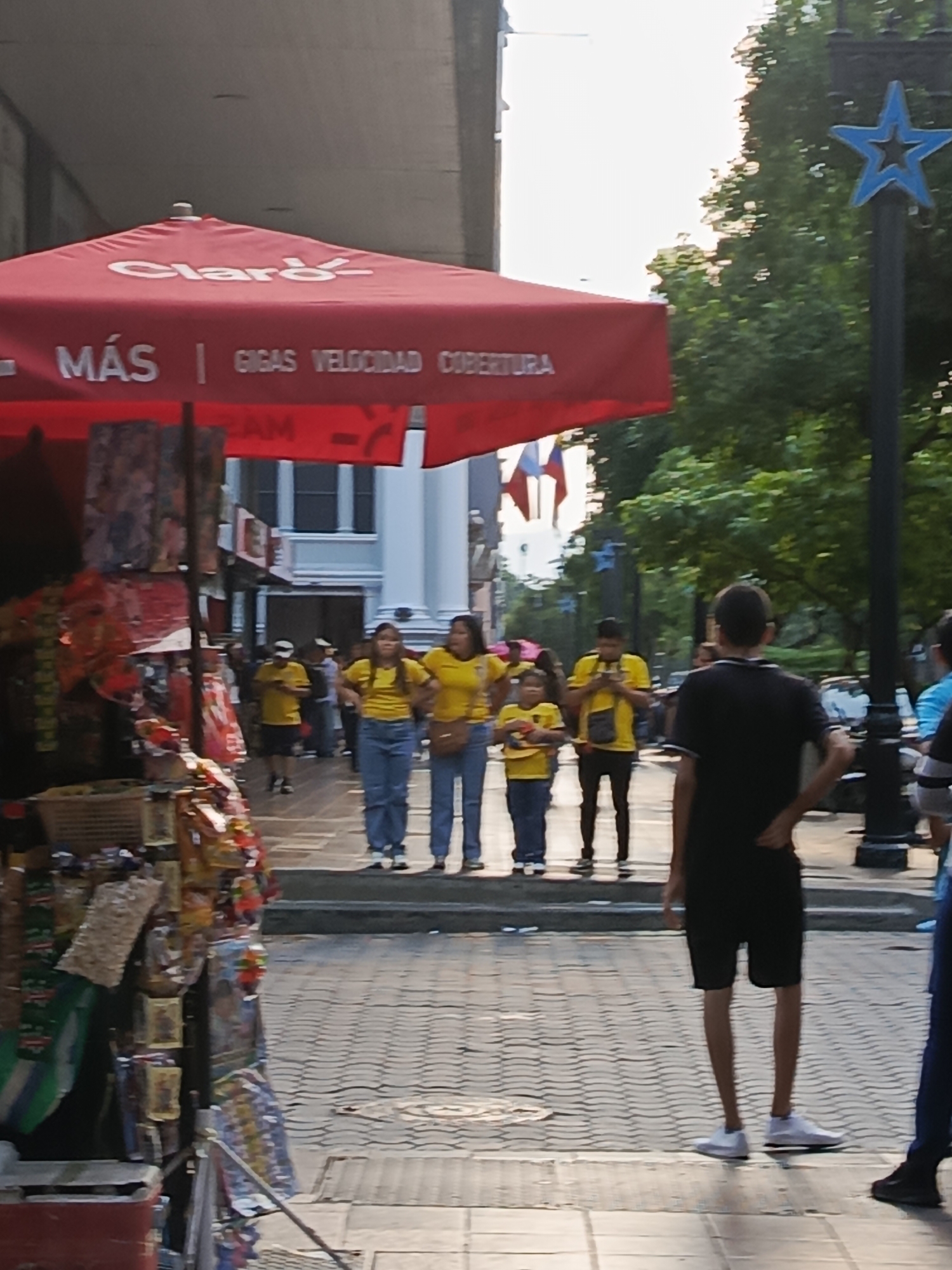 The image depicts a street scene in Guayaquil, Ecuador, possibly near a commercial area. A large red umbrella with "Claro" branding is prominent in the foreground, likely for an outdoor vendor. The umbrella advertises "MÁS | GIGAS VELOCIDAD COBERTURA".
Several individuals are visible on the sidewalk. Two women wearing yellow shirts and blue jeans stand together, facing towards the left of the frame. Behind them, a group of people in yellow shirts also appear to be standing or walking. One young child in a yellow shirt is also present. A person in a black t-shirt and shorts stands with their back to the camera, facing away from the umbrella.
The environment shows a paved sidewalk with a patterned design. In the background, there are buildings and trees. A black lamppost with a blue star-shaped decoration is visible to the right of the frame. The overall lighting suggests it is daytime. The presence of a street stall with various goods further suggests a commercial setting.