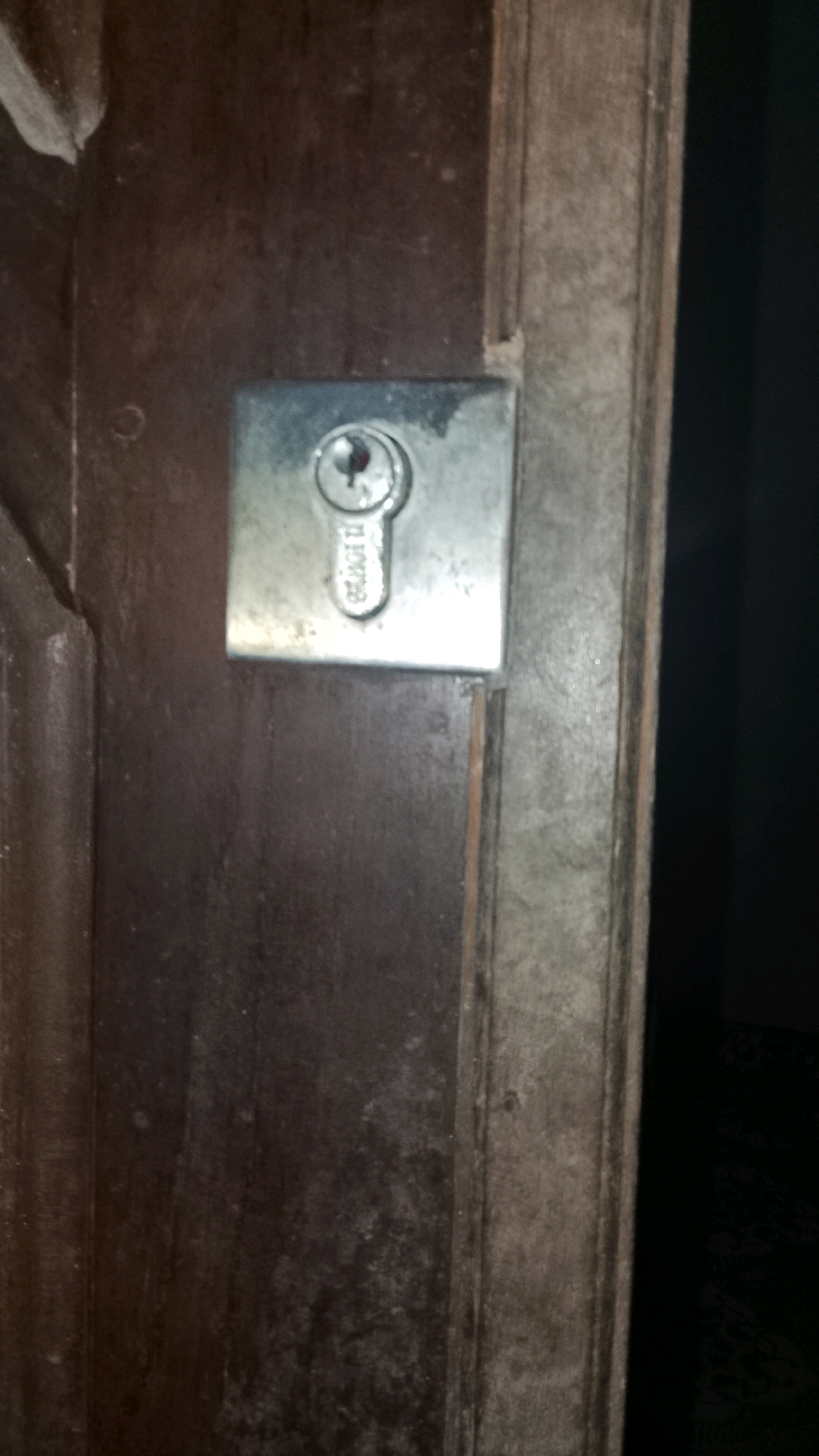 A close-up, eye-level shot captures a metal lock mechanism embedded in a dark, weathered wooden door. The lock, a dull silver color, features a circular keyhole with a partially visible key partially inserted. The surrounding doorframe appears to be made of a lighter, possibly painted or aged wood, with a rougher texture. The lighting is dim, casting shadows and highlighting the textures of the materials. The overall scene suggests an outdoor or slightly neglected environment, with no specific landmarks visible to indicate a precise location within Mulyo Haji, Indonesia.