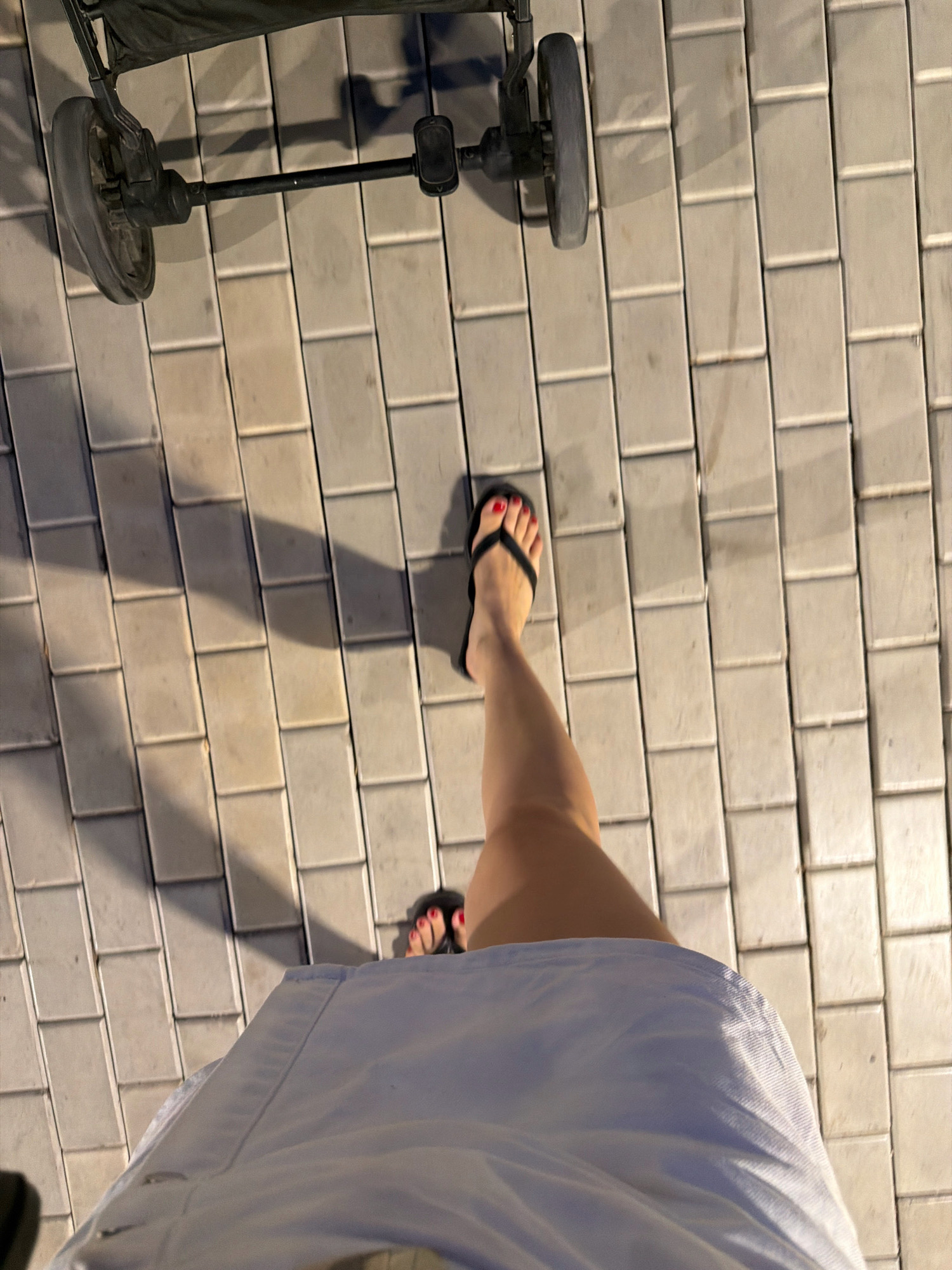 The image shows a person's legs and feet wearing black flip-flops with red nail polish, walking on a cobblestone path. A stroller is visible in the upper left corner of the image. The scene appears to be outdoors, possibly in a pedestrian area of Sharm El Sheikh, Egypt, given the presence of cobblestones and a stroller. Shadows cast by the stroller and the person indicate it is daytime, likely late afternoon or early evening. The overall atmosphere is casual and suggests a leisurely stroll.