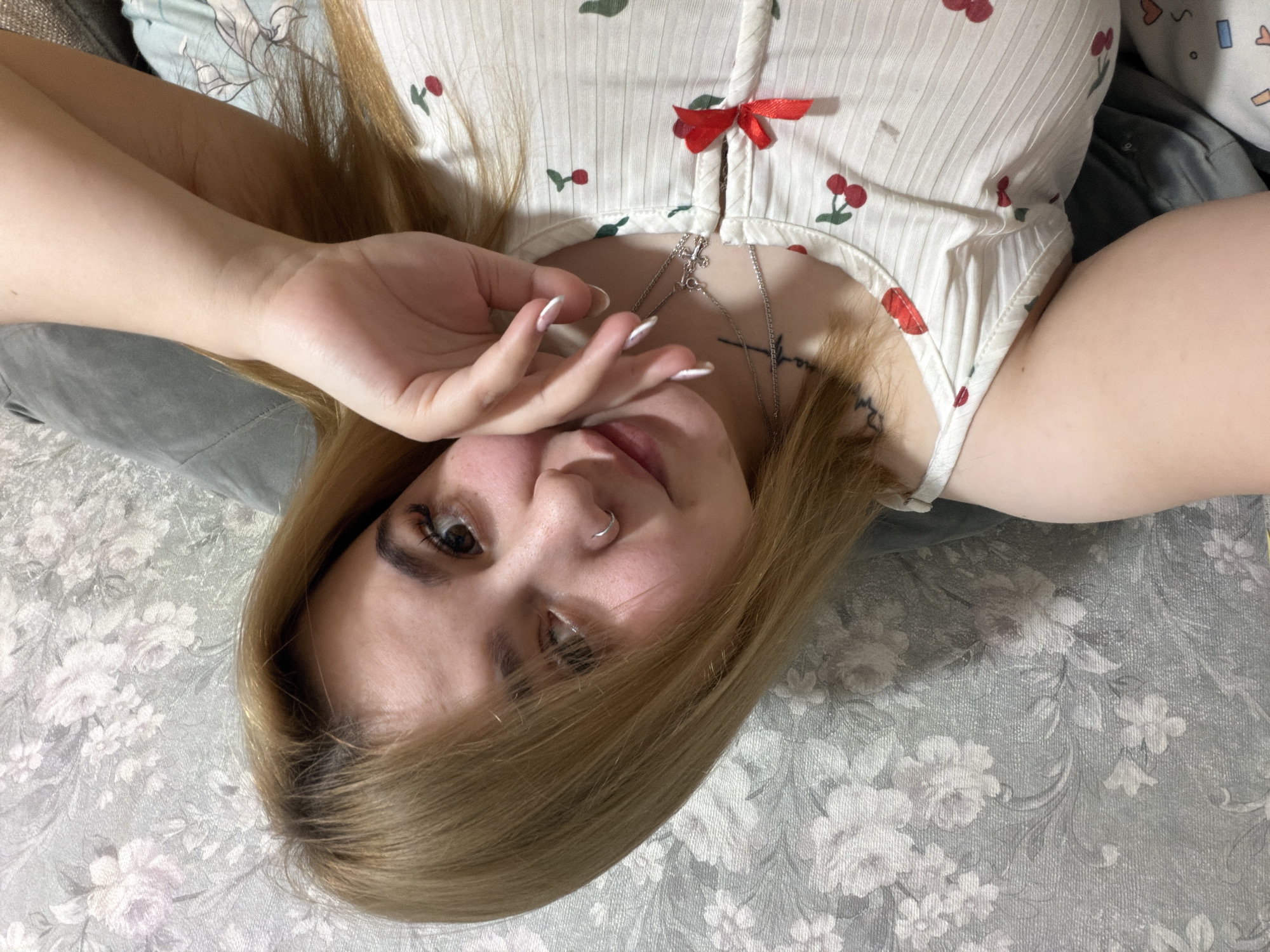 A young woman is lying on a bed with a floral patterned gray and white bedspread. She is wearing a white top with red cherry patterns and a small red bow. The woman has blonde hair and is partially covering her face with her hand. She has a silver nose ring and silver necklaces. The location cannot be determined from the image.