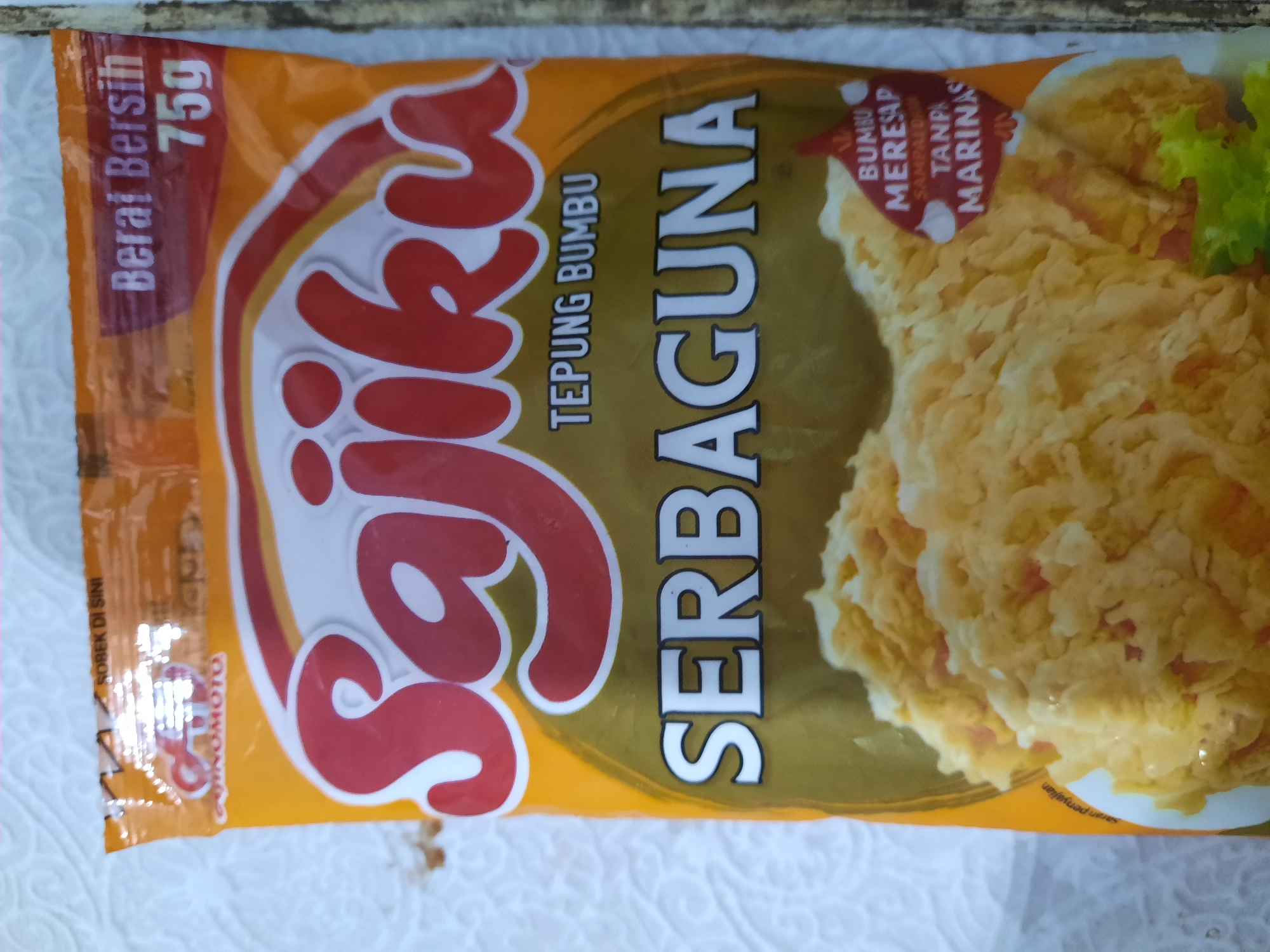 The image shows the front of a packet of "Sajiku Tepung Bumbu Serbaguna" (Sajiku All-Purpose Seasoned Flour). The packet is primarily orange and yellow, with prominent red text for the brand name "Sajiku." Below that, in white text, is "TEPUNG BUMBU" (Seasoned Flour) and "SERBAGUNA" (All-Purpose). The packet indicates a net weight of "75g." A close-up image of fried food, likely chicken, is displayed at the bottom. There is also a red label with white text that reads "BUMBU MEREAP SAMPAI DALAM TANPA MARINAS" (Seasoning Absorbs Deeply Without Marinating). The "AJI-NOMOTO" logo is visible in the upper left corner. The packet is placed on a white textured surface, possibly a tablecloth or paper. There is no discernible information in the image to suggest a location in Lhokseumawe, Indonesia.