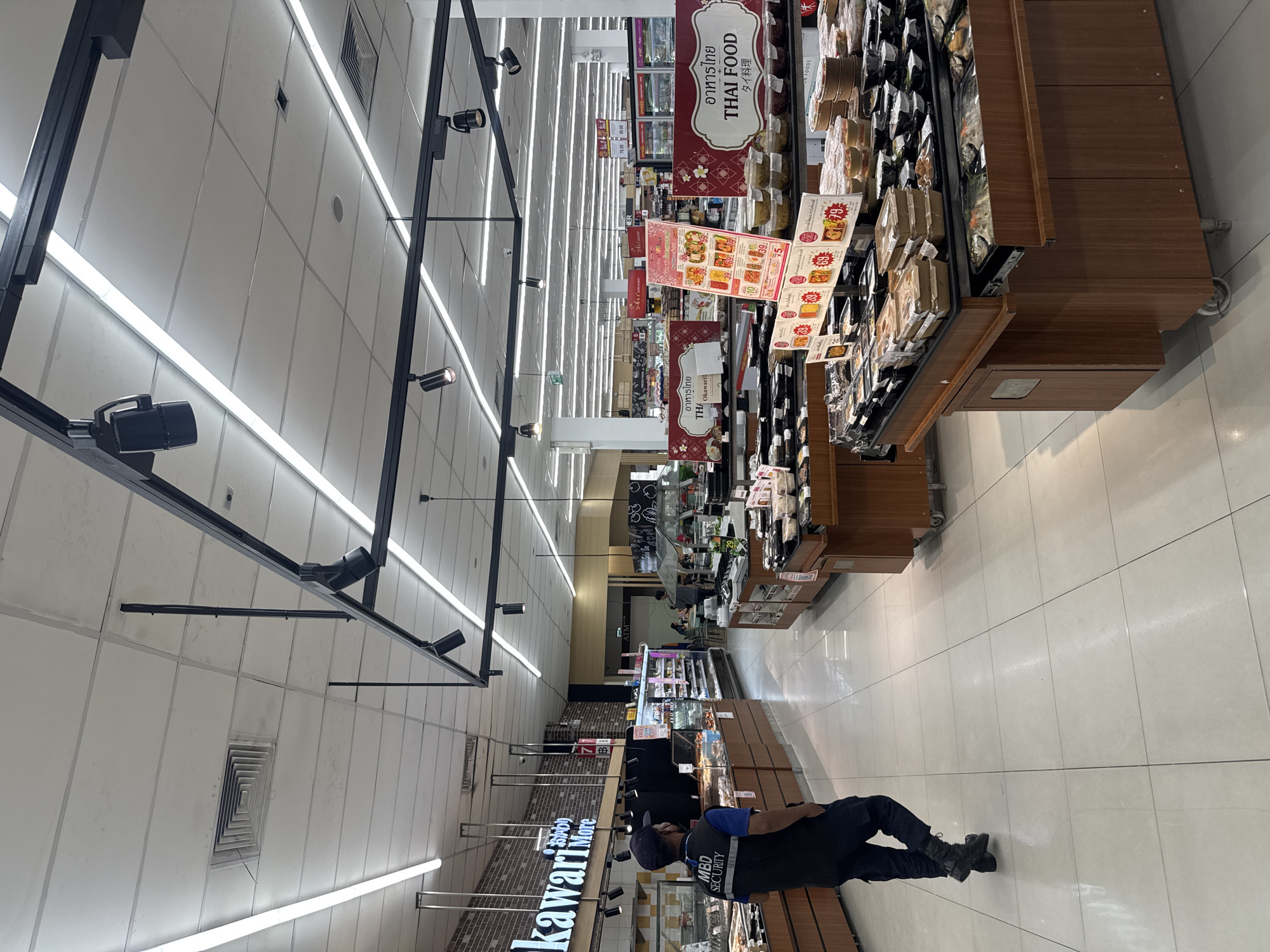 The image shows a security guard wearing a dark uniform with "MBD SECURITY" written on the vest, walking away from the camera inside a supermarket. The supermarket has tiled floors and a suspended ceiling with fluorescent lights and track lighting. In the background, there are aisles stocked with packaged food items, including what appears to be ready-to-eat meals and produce. Signage in Thai and English indicates "THAI FOOD" and "Okawari More." The overall impression is of a well-lit, modern grocery store. The specific location within Bangkok, Thailand cannot be determined from the image alone.