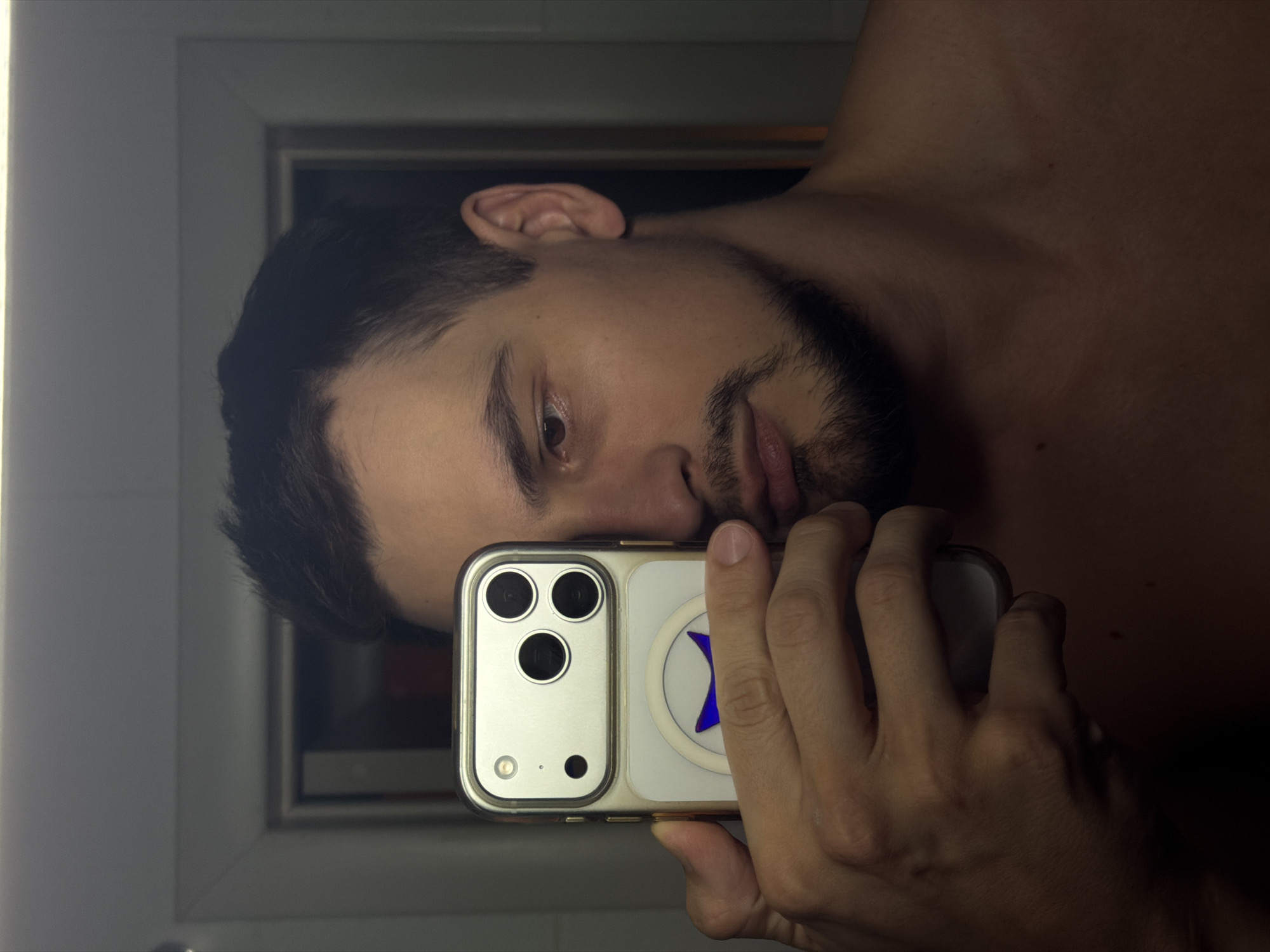 A man with dark hair and a trimmed beard is taking a selfie with a smartphone. The phone has a white case with a circular MagSafe accessory on the back. The man's bare shoulder is visible. The background shows a window frame. The location appears to be indoors. There is no information to suggest this image was taken in Krasnoyarsk, Russia.