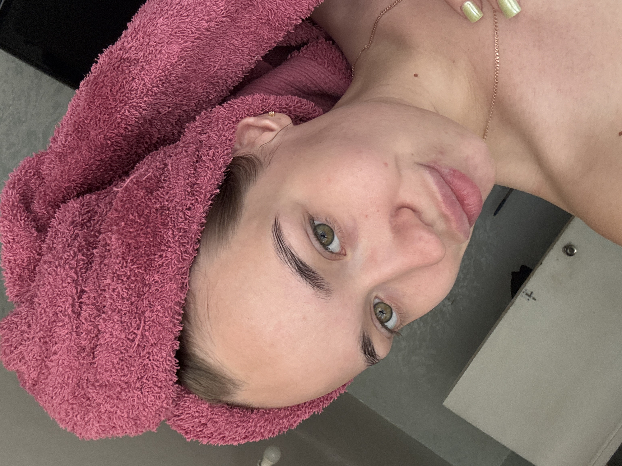 A young woman with fair skin, light brown hair, and green eyes is seen from an upward angle. She is wearing a pink towel wrapped around her head and has a gold necklace. Her fingernails are painted a light green. The background is a neutral gray, with a white cabinet visible in the lower right. It is unclear where exactly in Chaikovsky, Russia this image was taken.