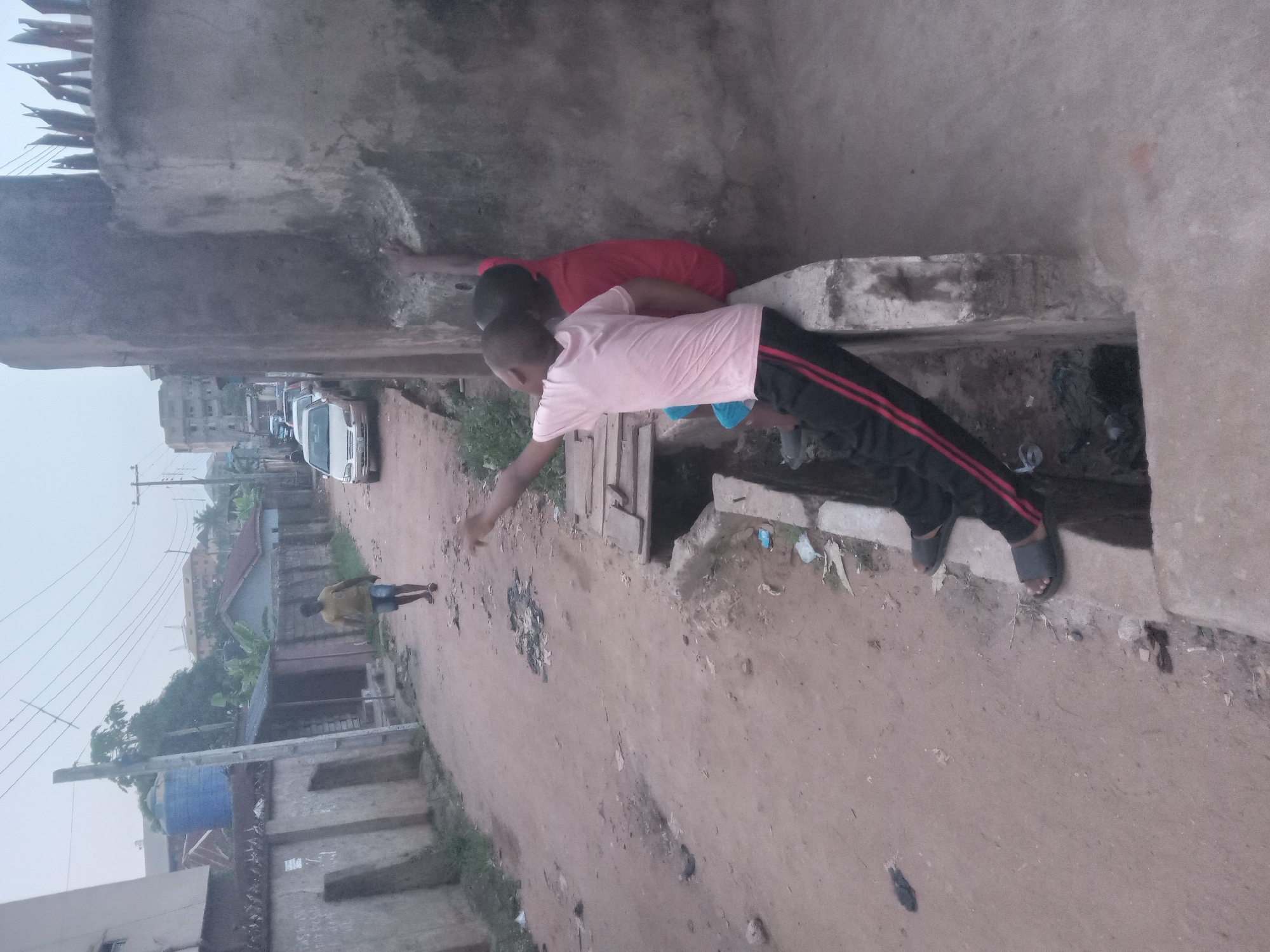 Two boys are positioned on a concrete structure beside a dirt road in Nkpor, Nigeria. One boy wears a pink shirt and dark pants with red stripes, while the other wears a red shirt. The boy in pink reaches toward the road with his arm extended. In the background, there are houses, trees, and a person walking along the road. Several cars are parked further up the road. The weather appears overcast.